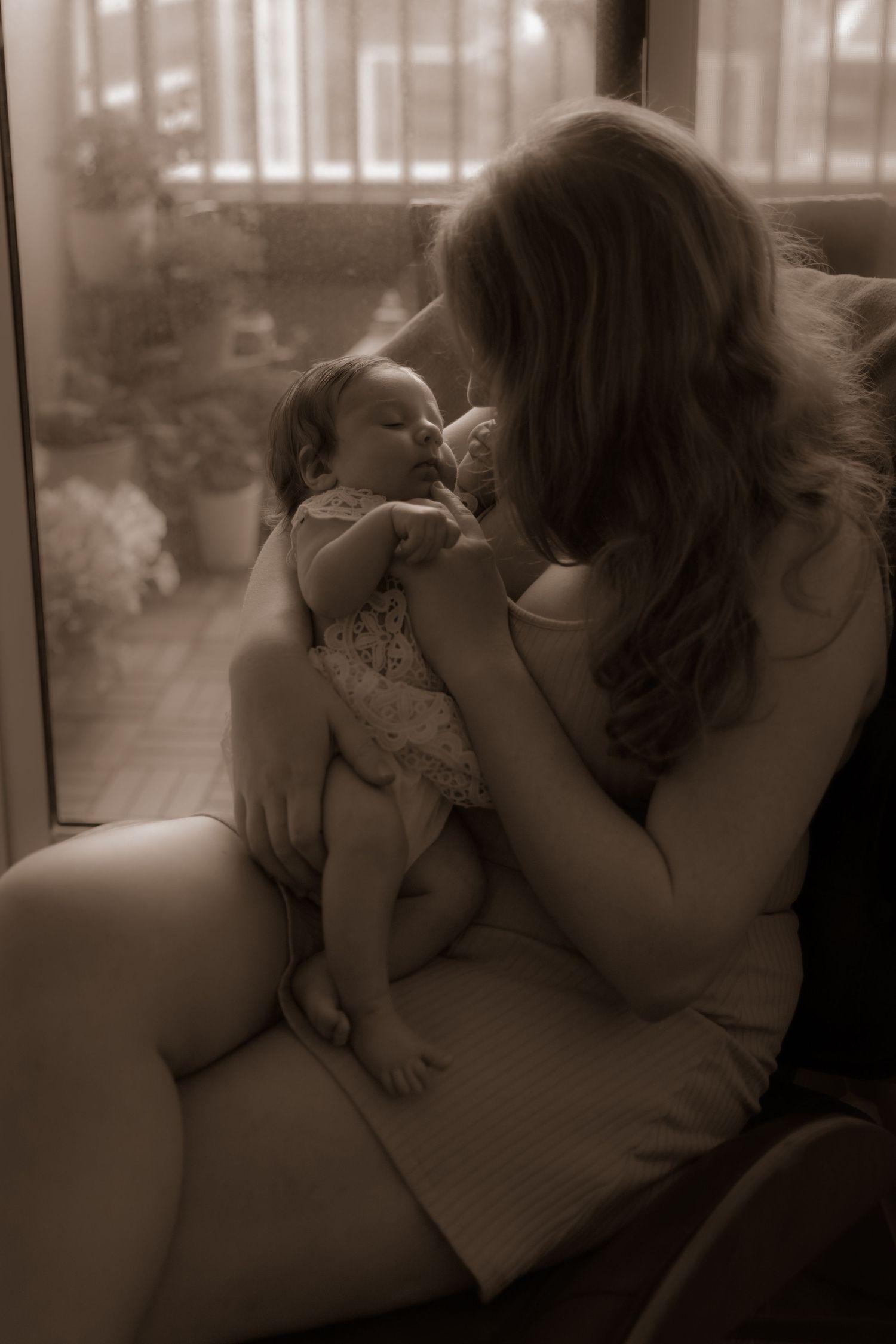 A tender sepia-toned portrait of a parent cradling a newborn baby in their arms by a window.