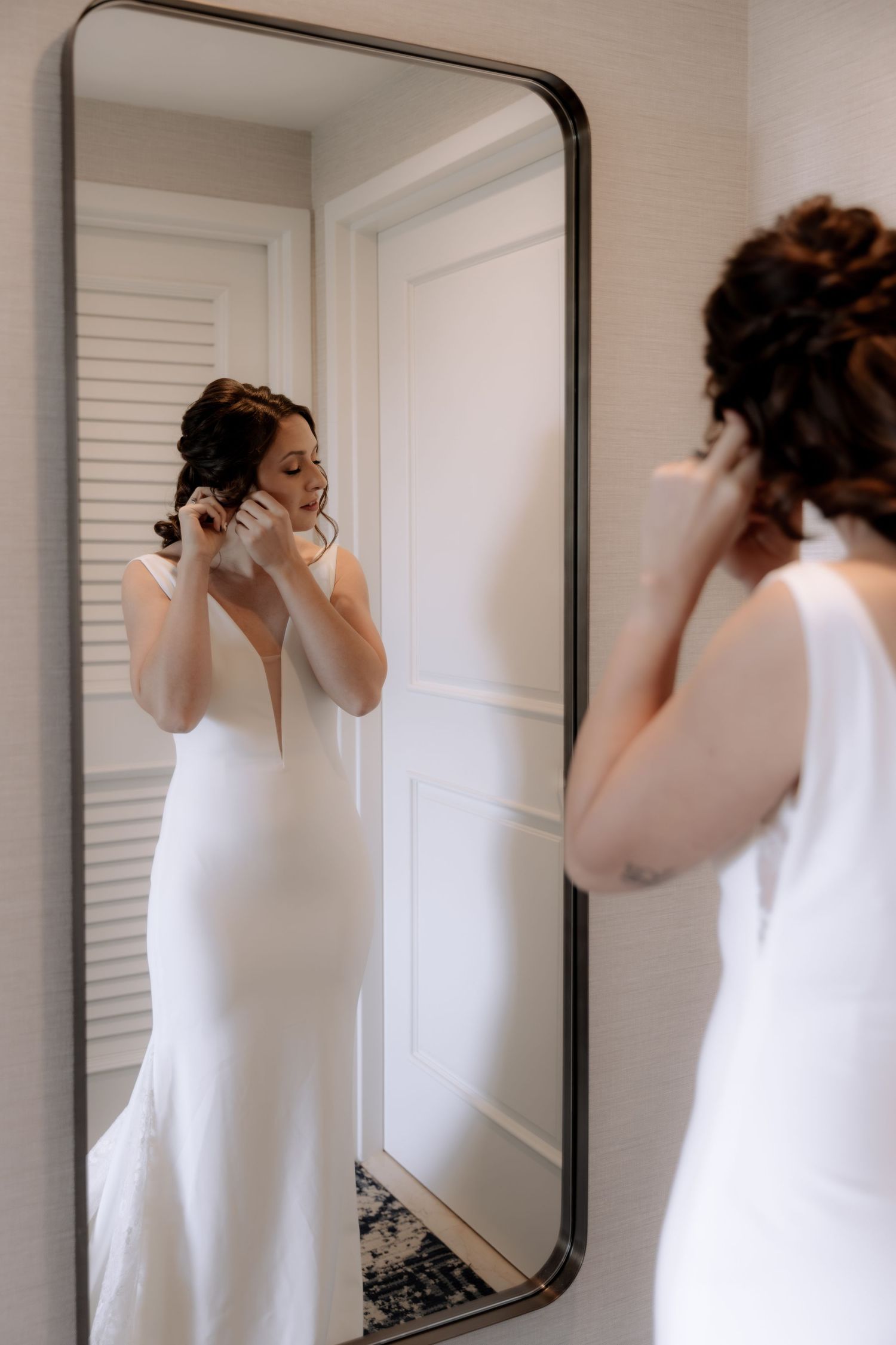 A bride in an elegant white wedding dress adjusts her earrings while looking in a full-length mirror.