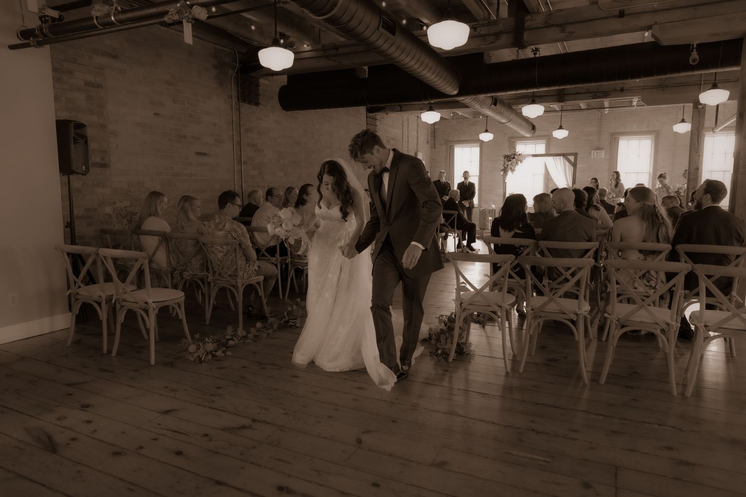A sepia wedding photograph showing a romantic processional through a venue with warm lighting and brick walls.