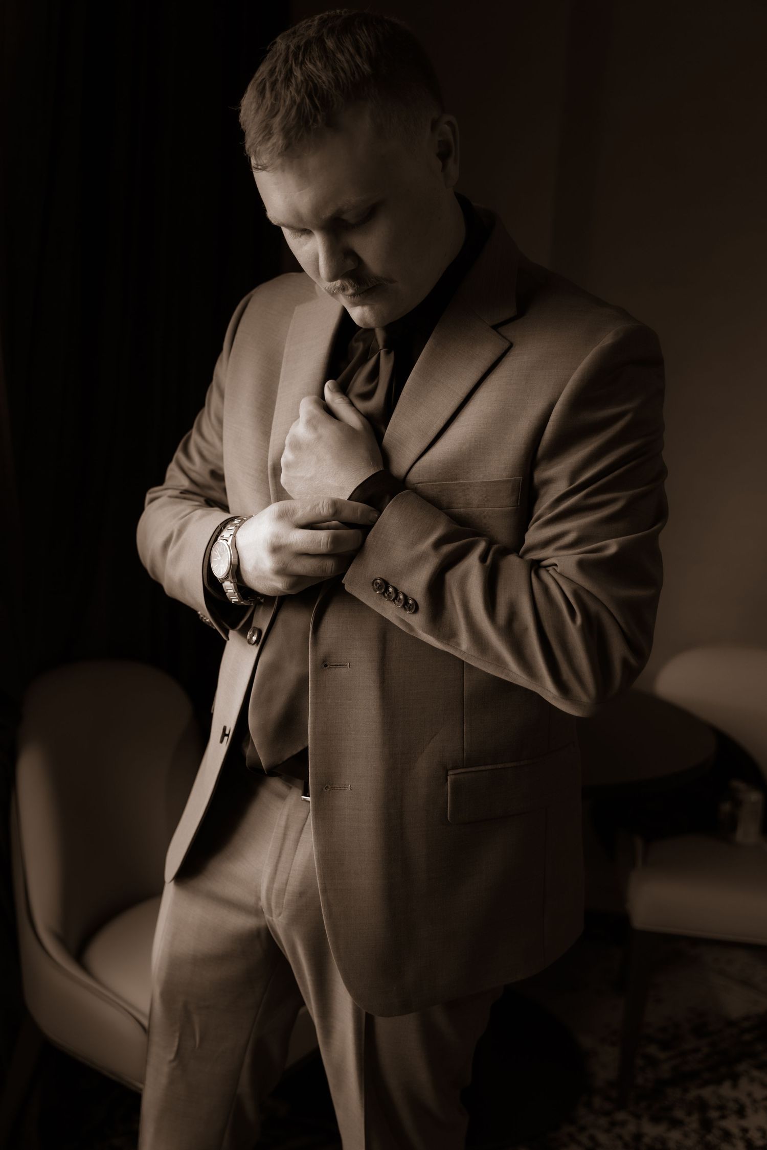 A figure in a tan suit adjusts their tie while standing in a dimly lit room with moody lighting.