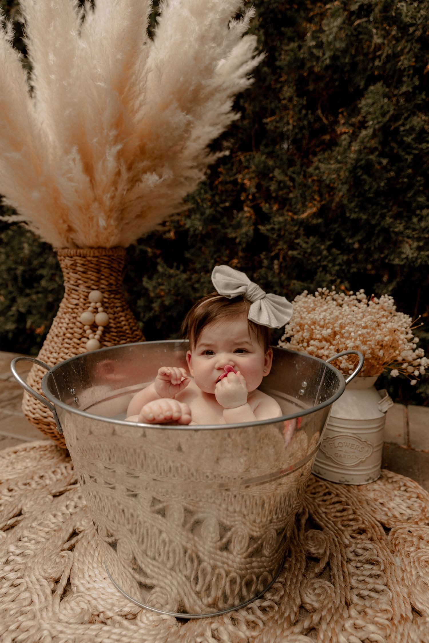 Baby taking milk bath in ornate metal tub with pampas grass and boho decor in outdoor setting.