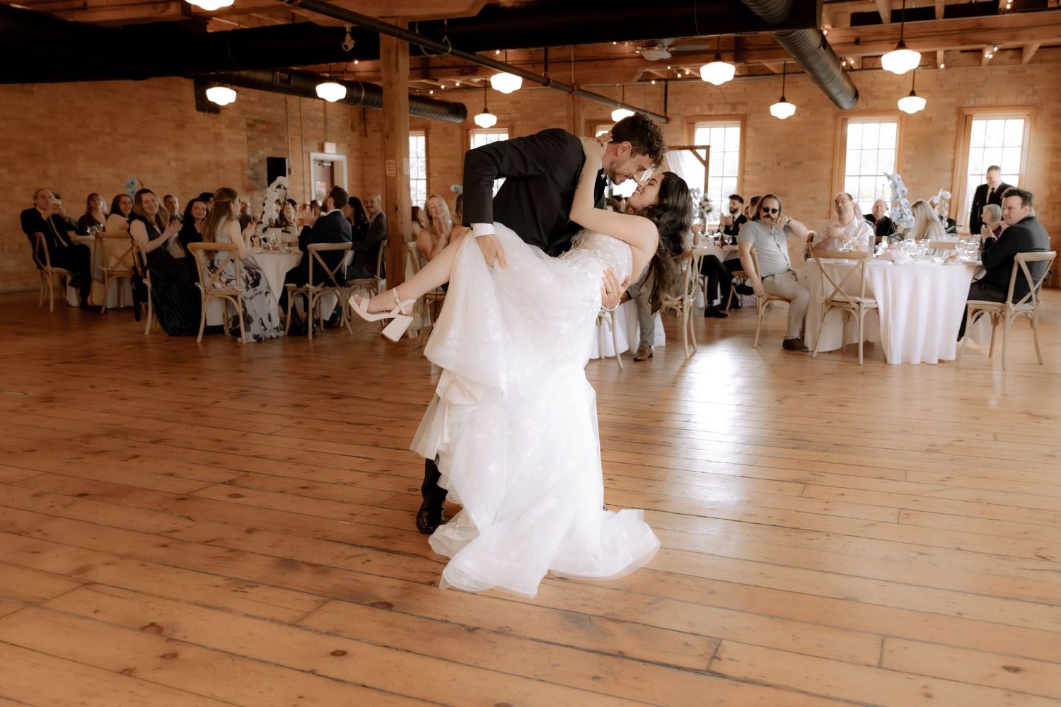 A couple shares their first dance at a wedding reception in a rustic venue with wooden floors and pendant lighting.