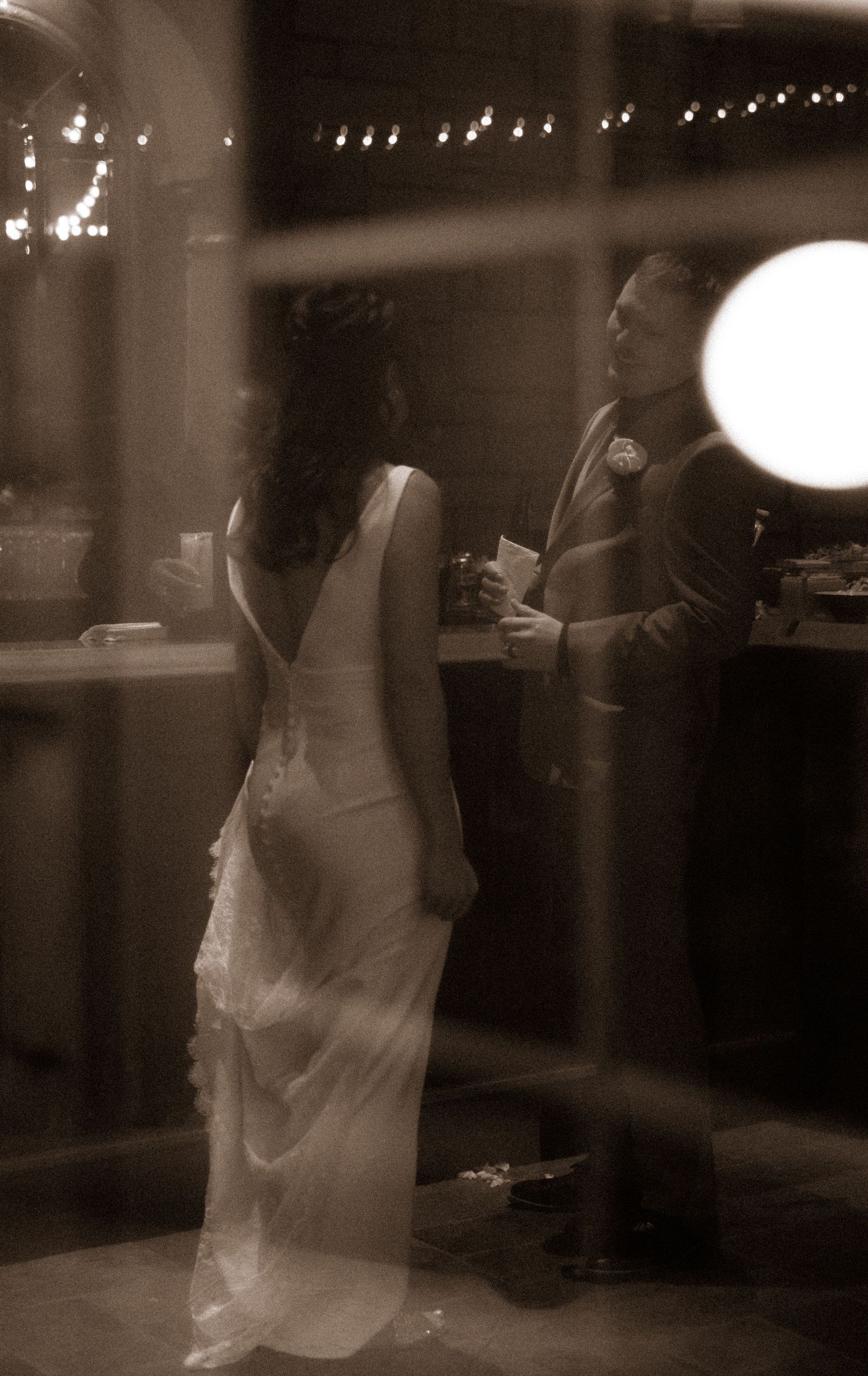 Soft-focus view through window of couple sharing conversation at wedding reception bar.