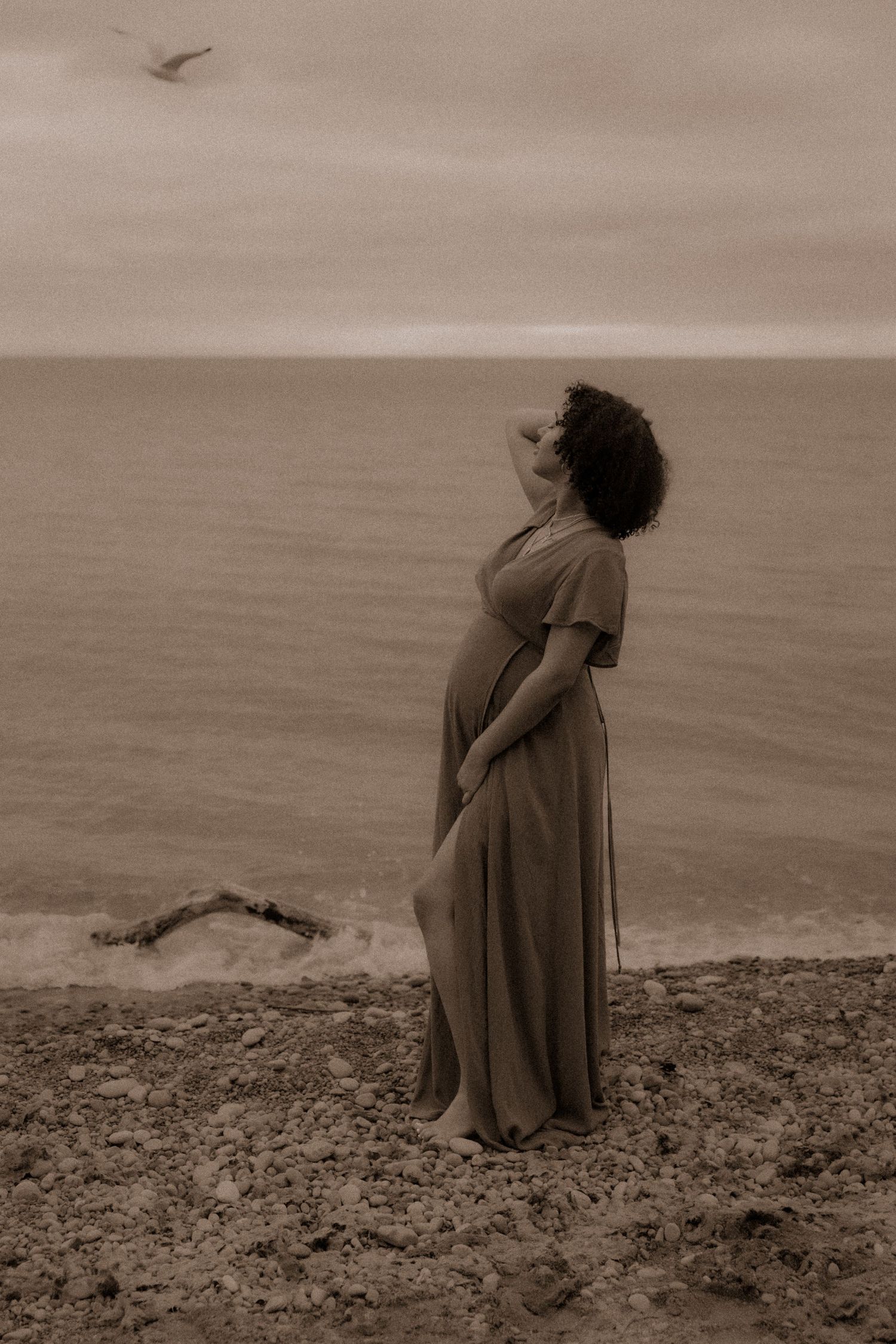 A maternity photoshoot shows an expectant woman in a flowy dress standing on a beach in sepia tones.