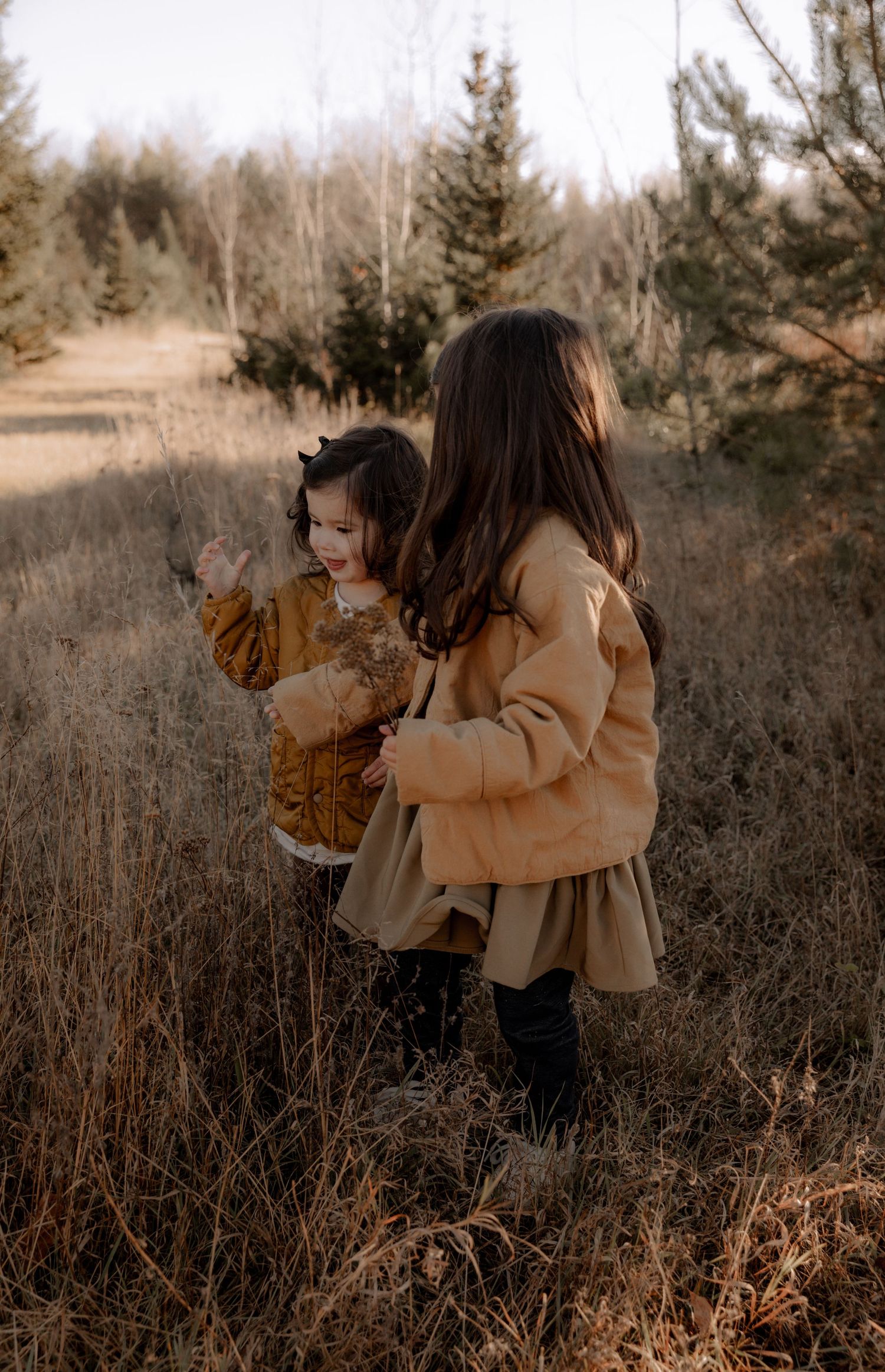 Two people in matching camel-colored coats explore a winter landscape with dried grass and evergreens.