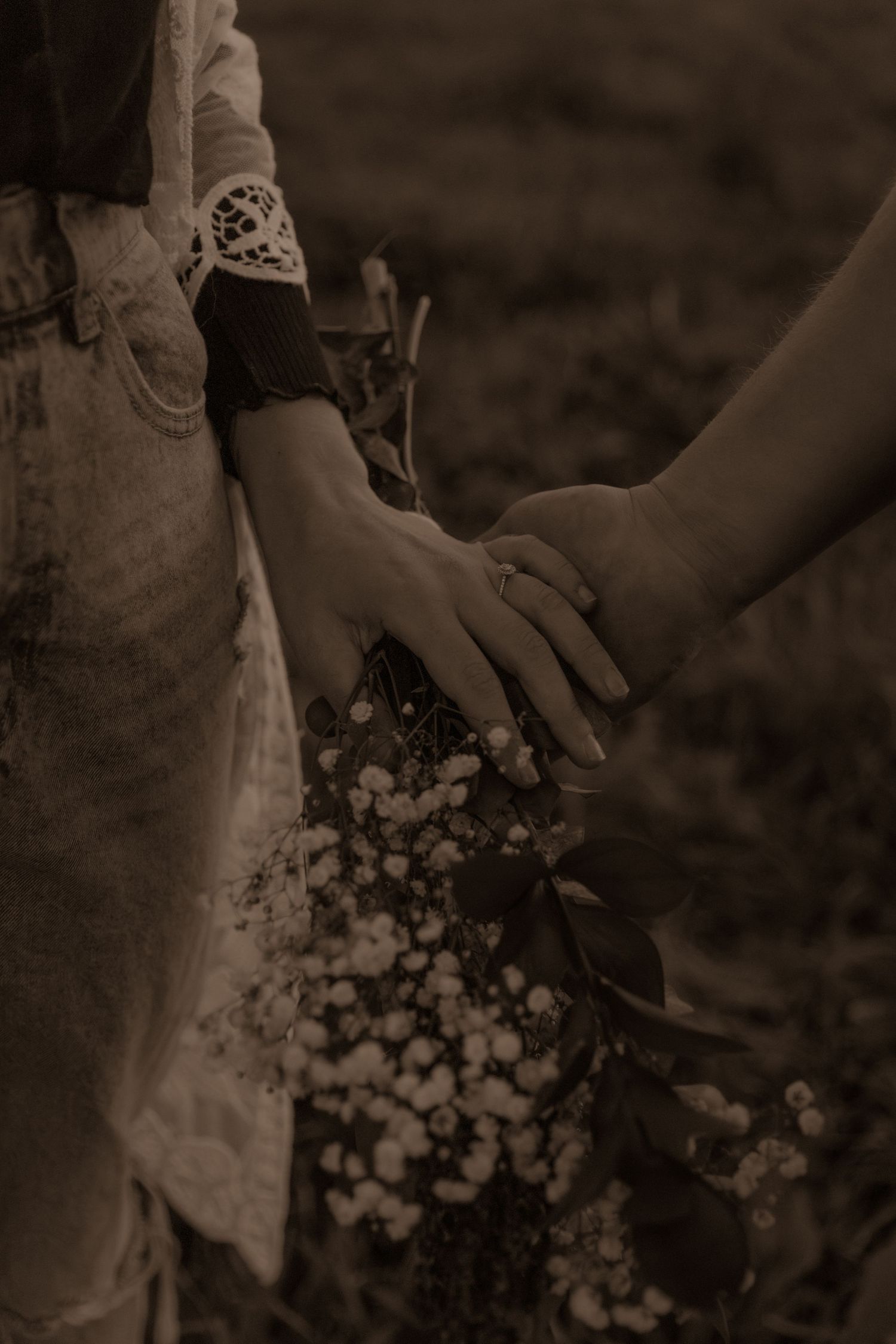Sepia-toned image of hands holding with lace sleeve and delicate flowers.