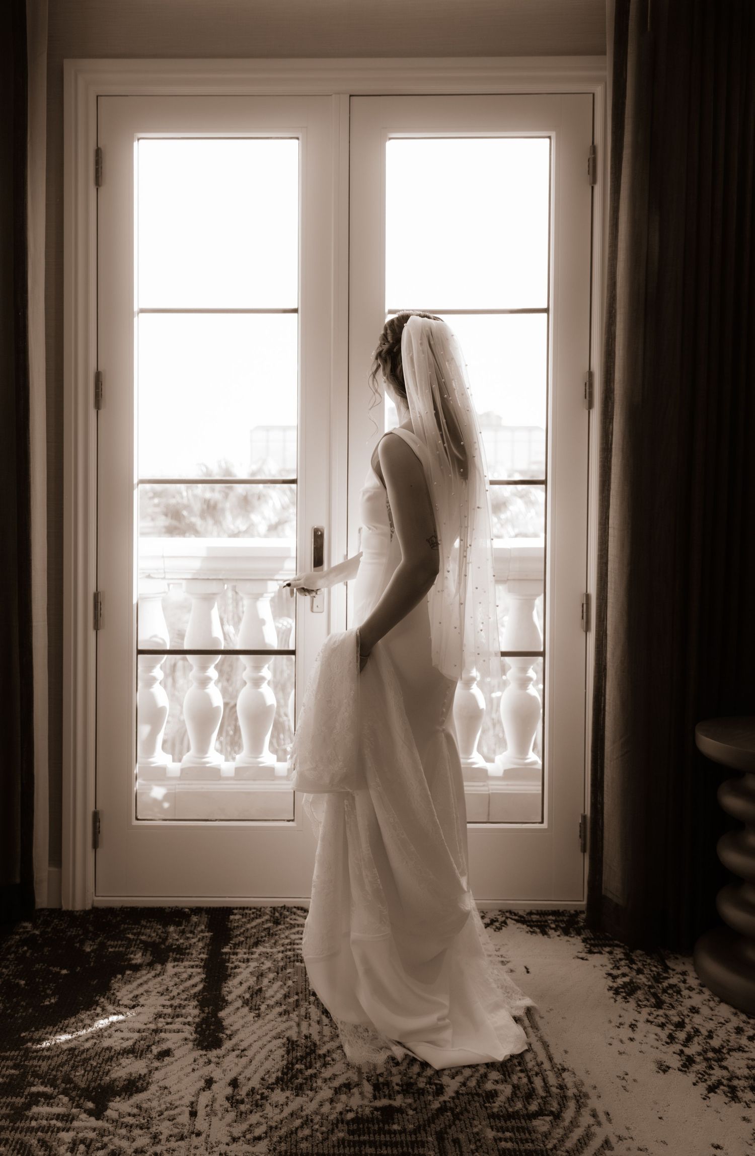 An elegant bridal silhouette posed against bright French doors with ornate balustrade details.