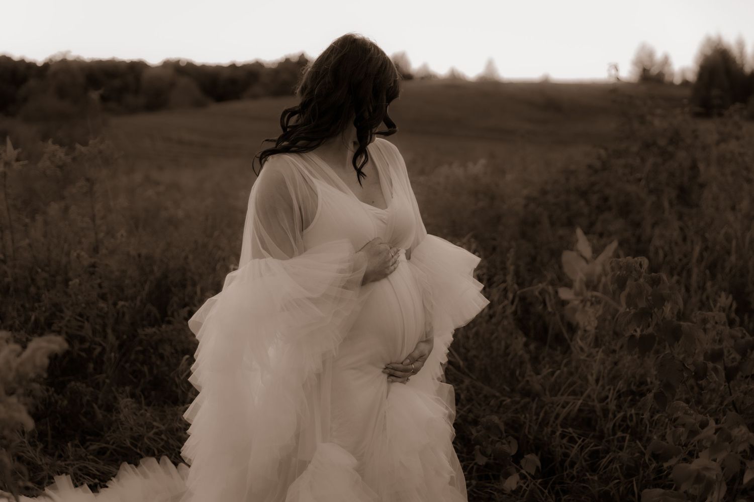 An artistic maternity portrait in sepia tones showcasing a flowing white dress in nature.