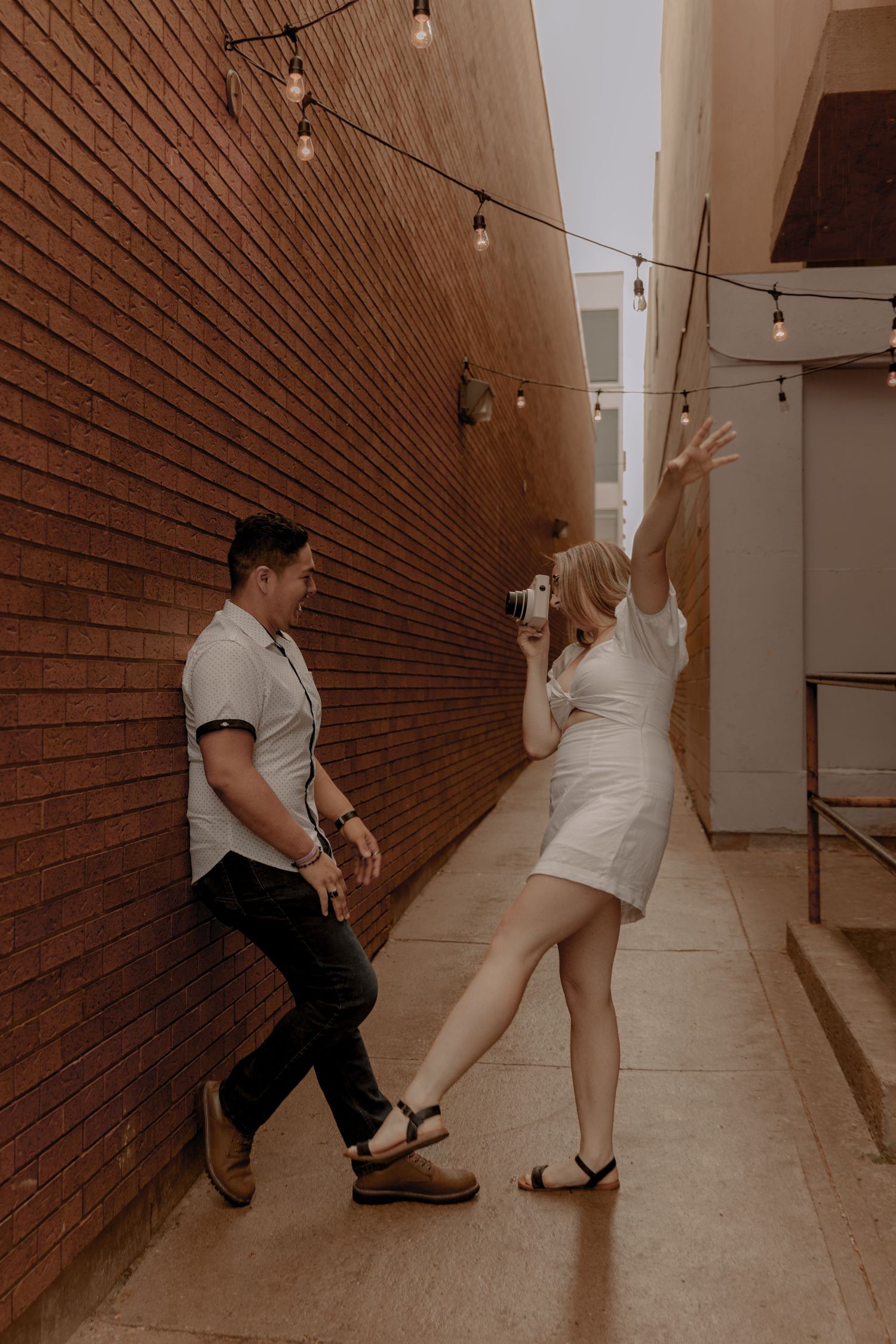Two people enjoying a playful moment in an urban alley with string lights overhead and brick walls.