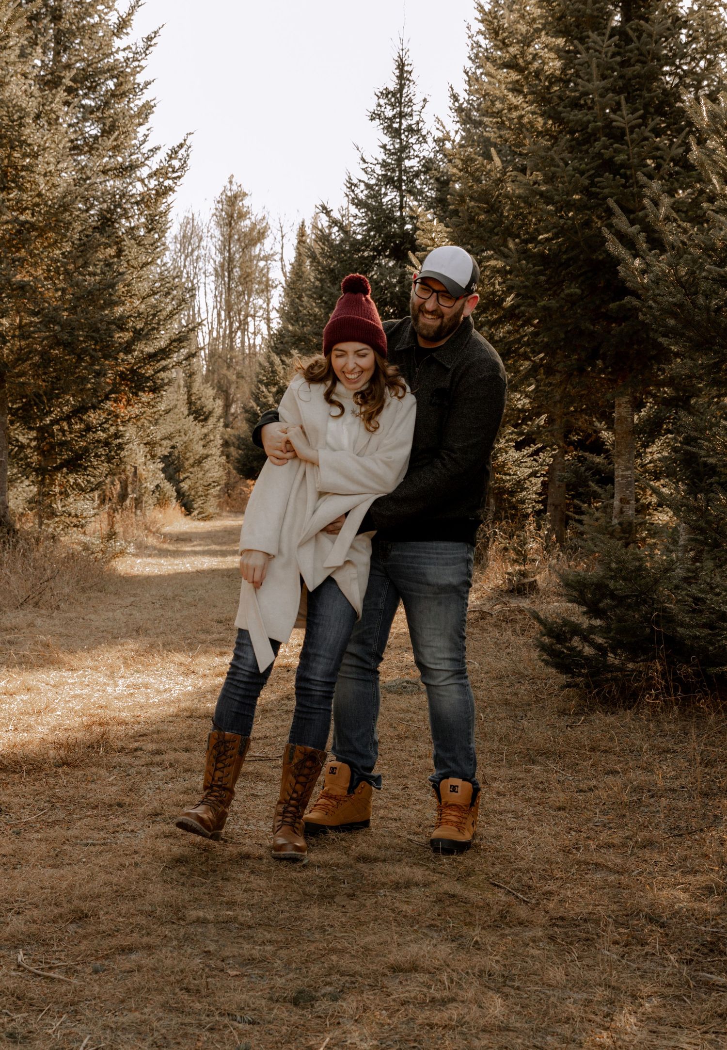 A couple embraces on a forest trail in autumn wearing cozy winter clothing and boots against an evergreen backdrop.