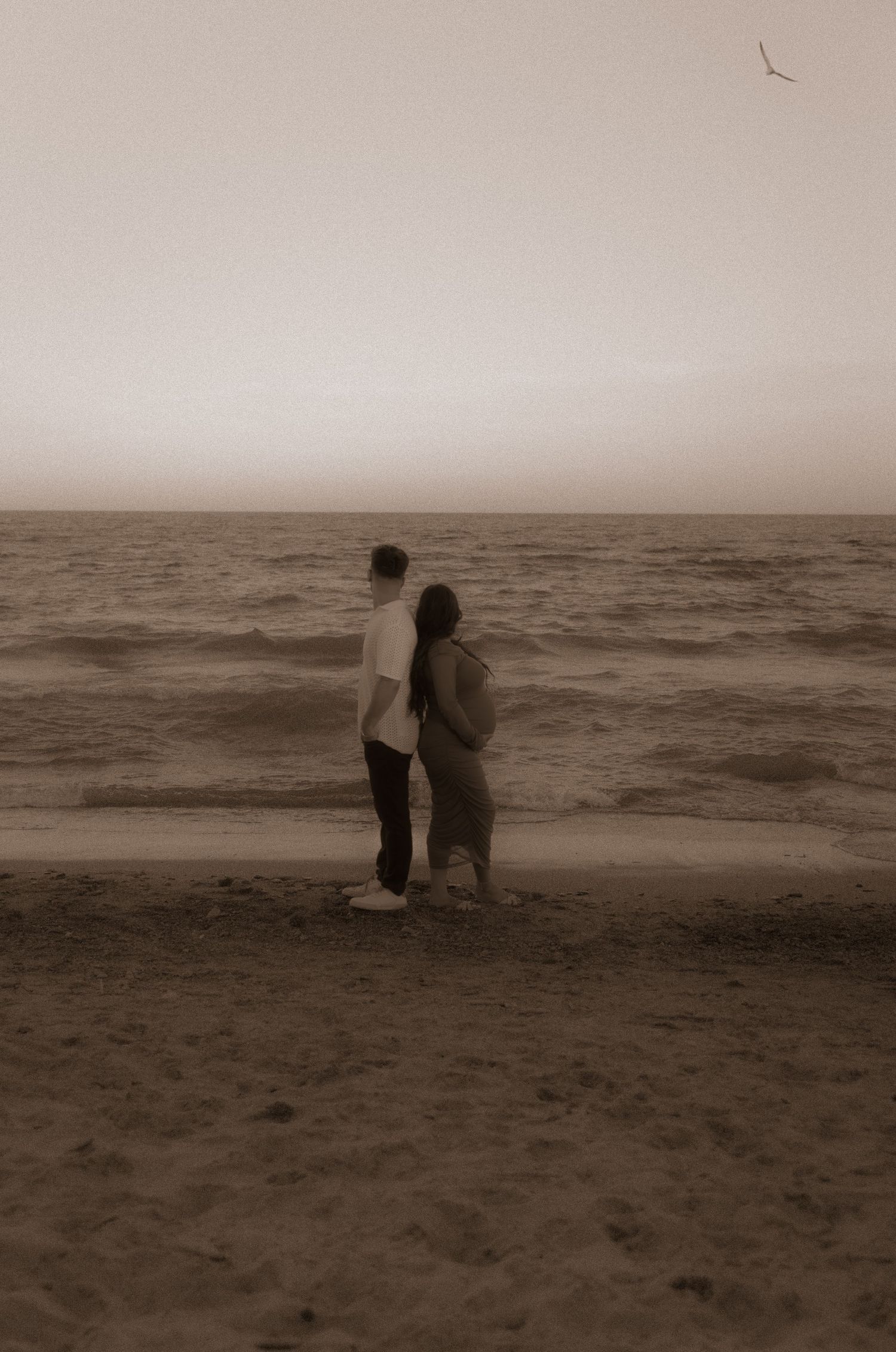 Two silhouetted figures stand together at the edge of a misty beach shoreline in sepia tones at dusk.