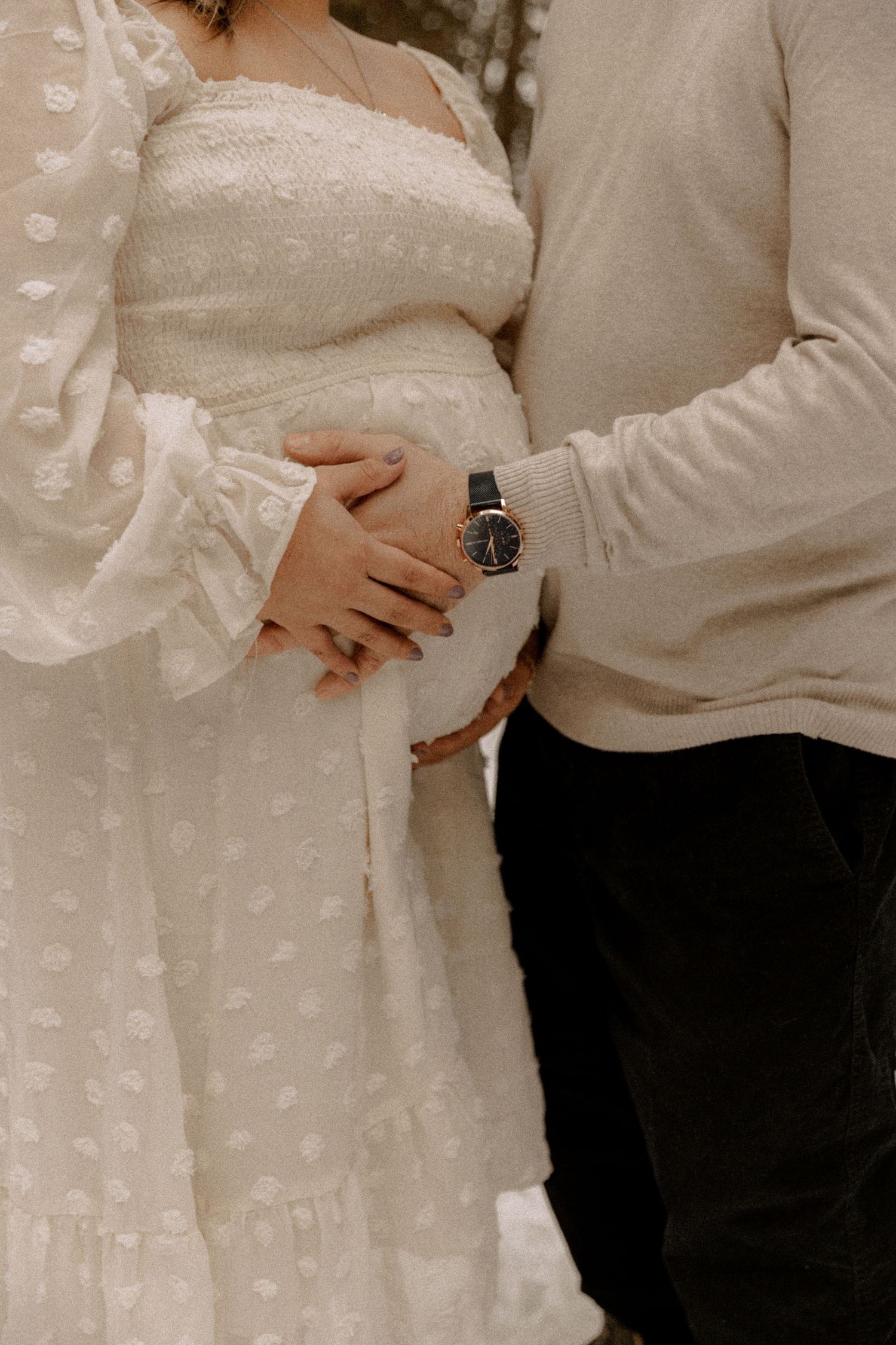 A tender maternity moment showing hands resting on a pregnant belly, wearing a white lace dress beside someone in neutral tones.