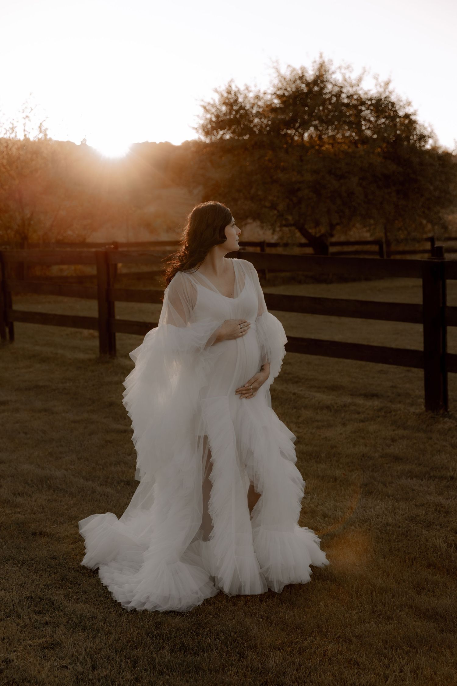 A dreamy maternity photoshoot in a flowing white gown at sunset with wooden fences and trees in the background.