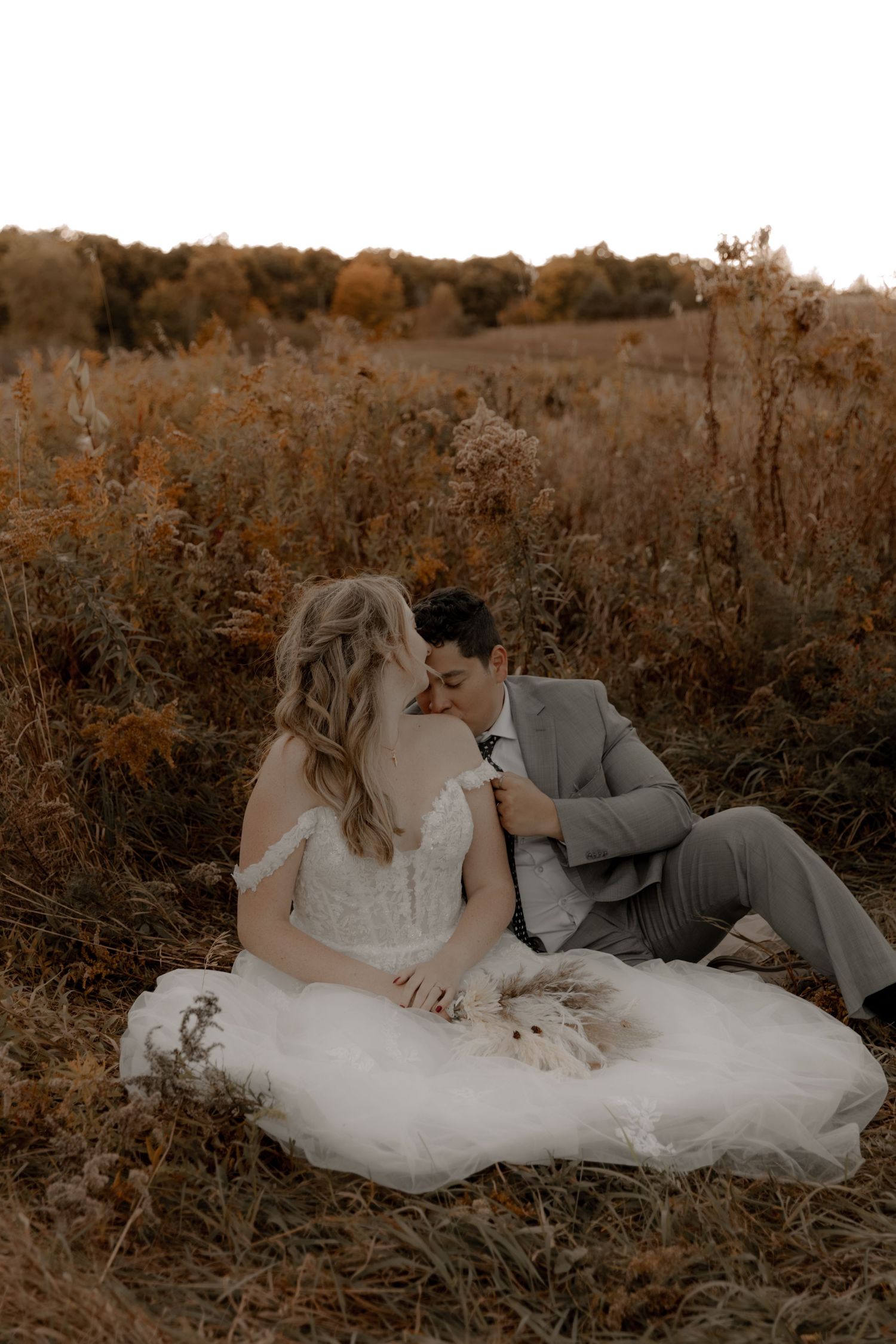 A romantic wedding portrait of a couple sitting in tall autumn grass at sunset.