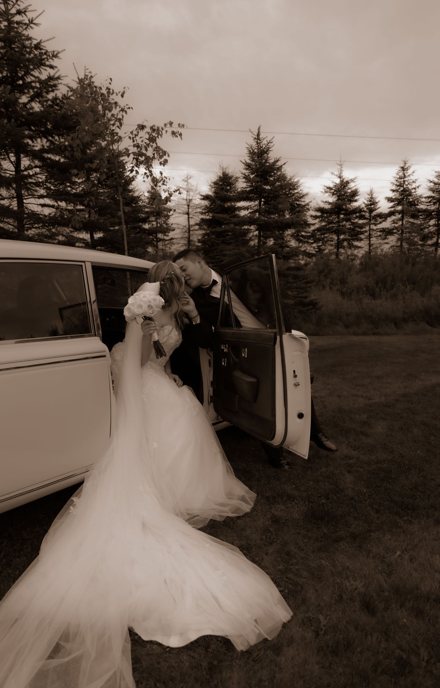 Romantic vintage wedding scene with flowing white dress and classic car among evergreen trees in sepia tones.