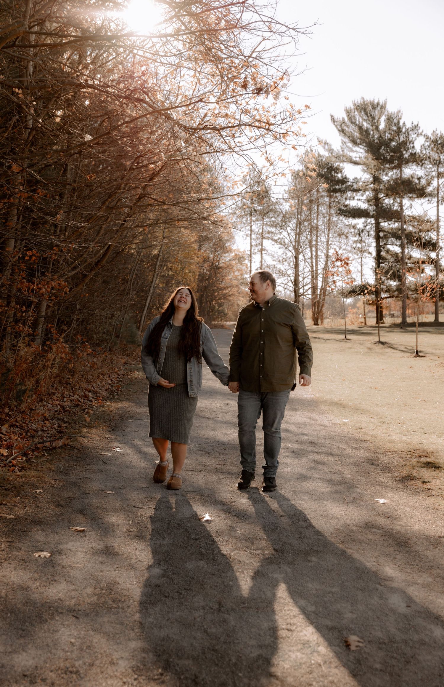 A couple walks together on an autumn path surrounded by bare trees casting long shadows on the ground.