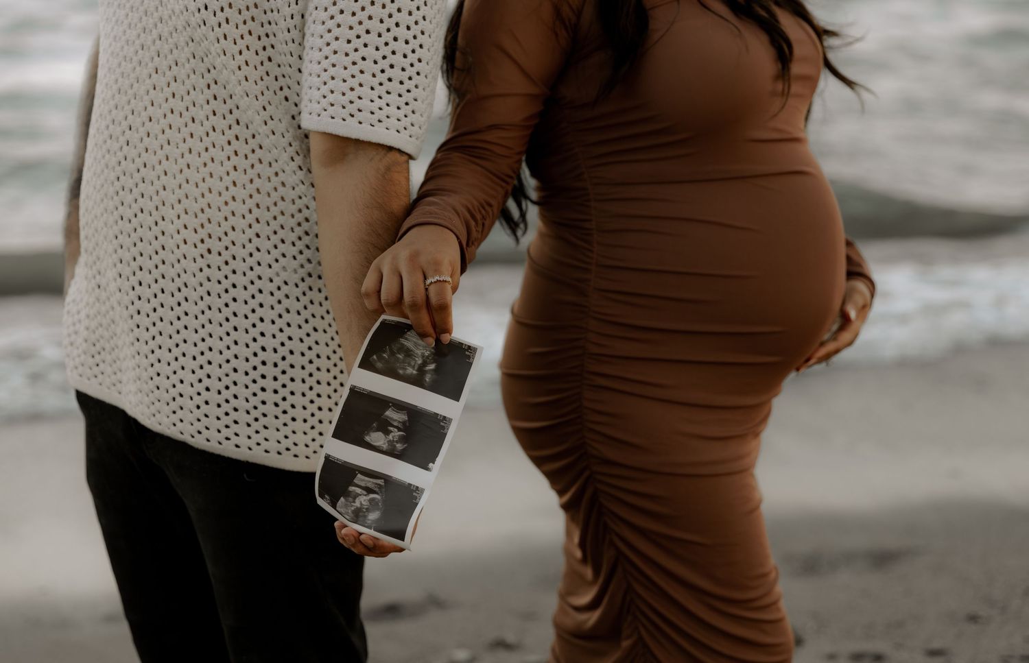 A couple at the beach hold ultrasound photos while wearing a white knit sweater and brown fitted maternity dress.