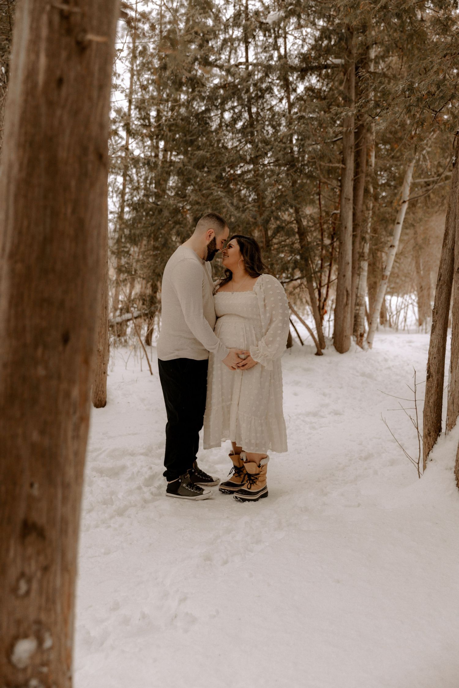 A couple standing close together in a snowy winter forest, surrounded by tall trees casting long shadows.