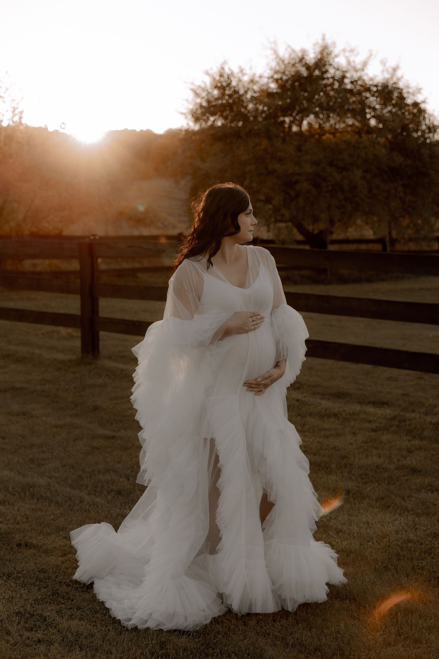 A dreamy maternity photo at sunset shows an expectant mother in a flowing white tulle gown against a fenced pasture backdrop.