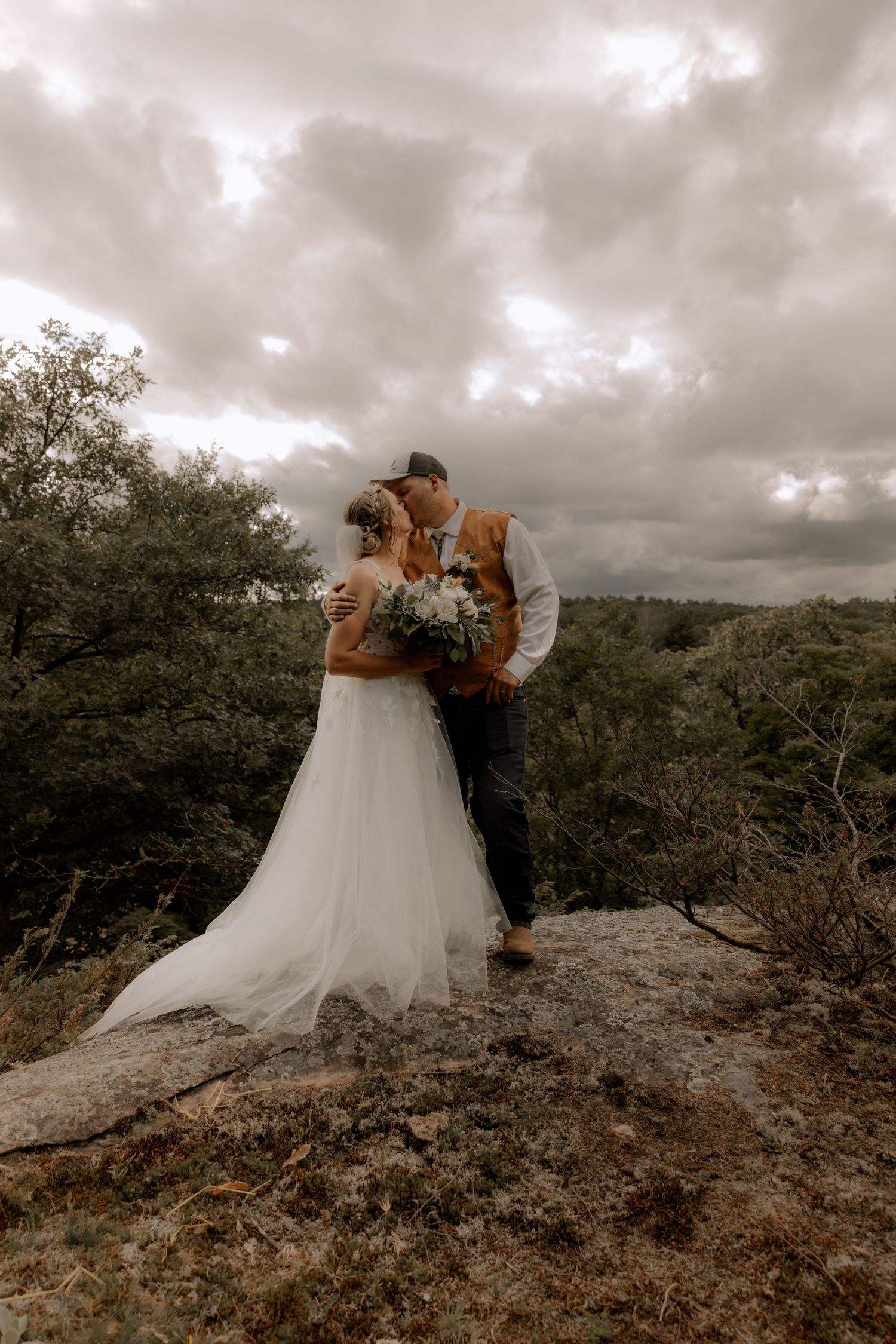 A dramatic wedding photo of a couple embracing on a rocky cliff under moody storm clouds.