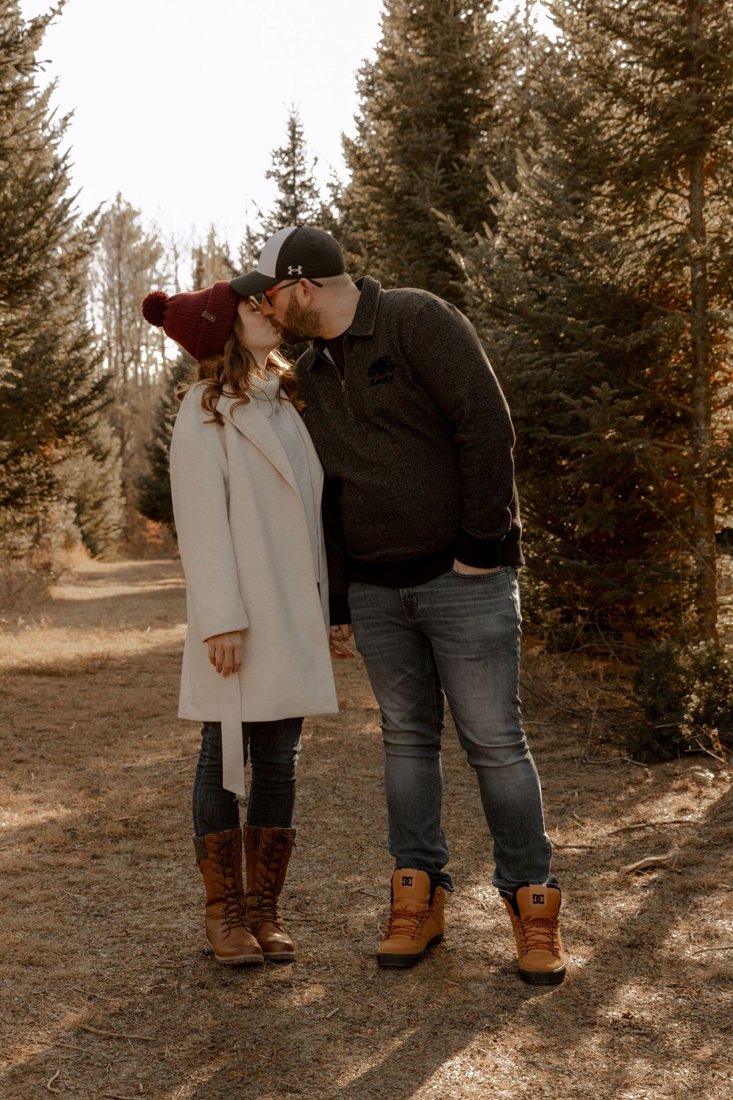 A couple sharing an intimate moment on a forest path in autumn wearing coats and boots against a backdrop of evergreen trees.