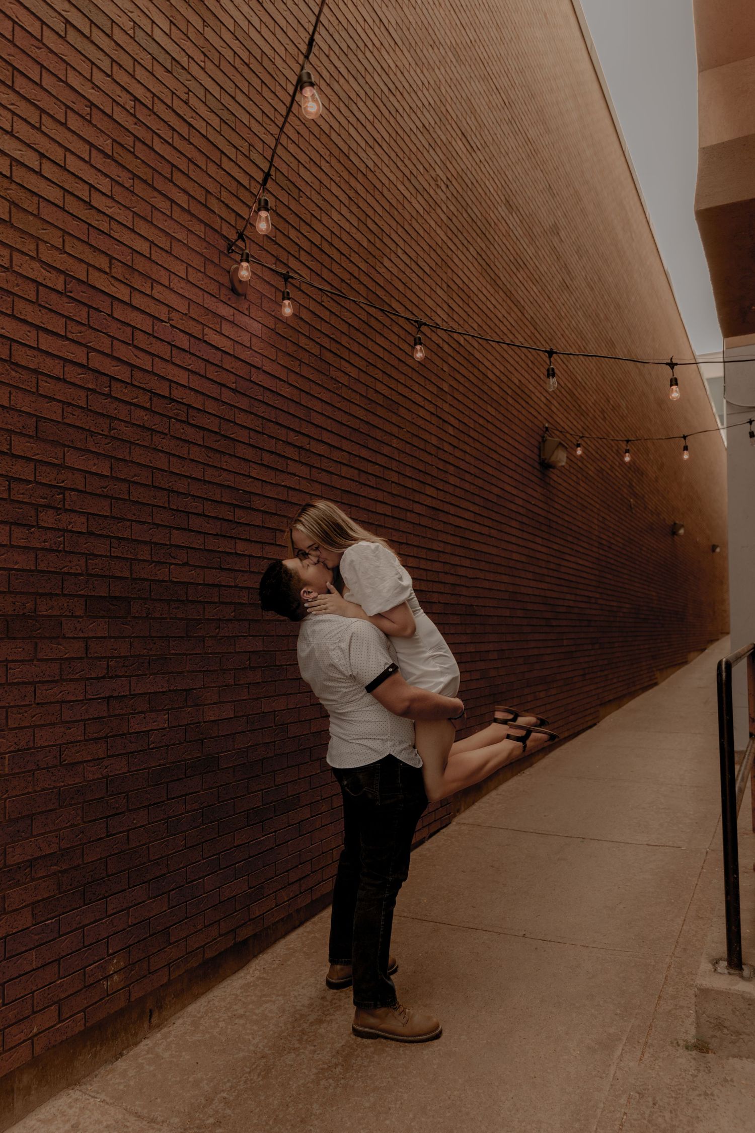 Silhouette of a couple in a romantic embrace against a brick wall with string lights in a dimly lit hallway.