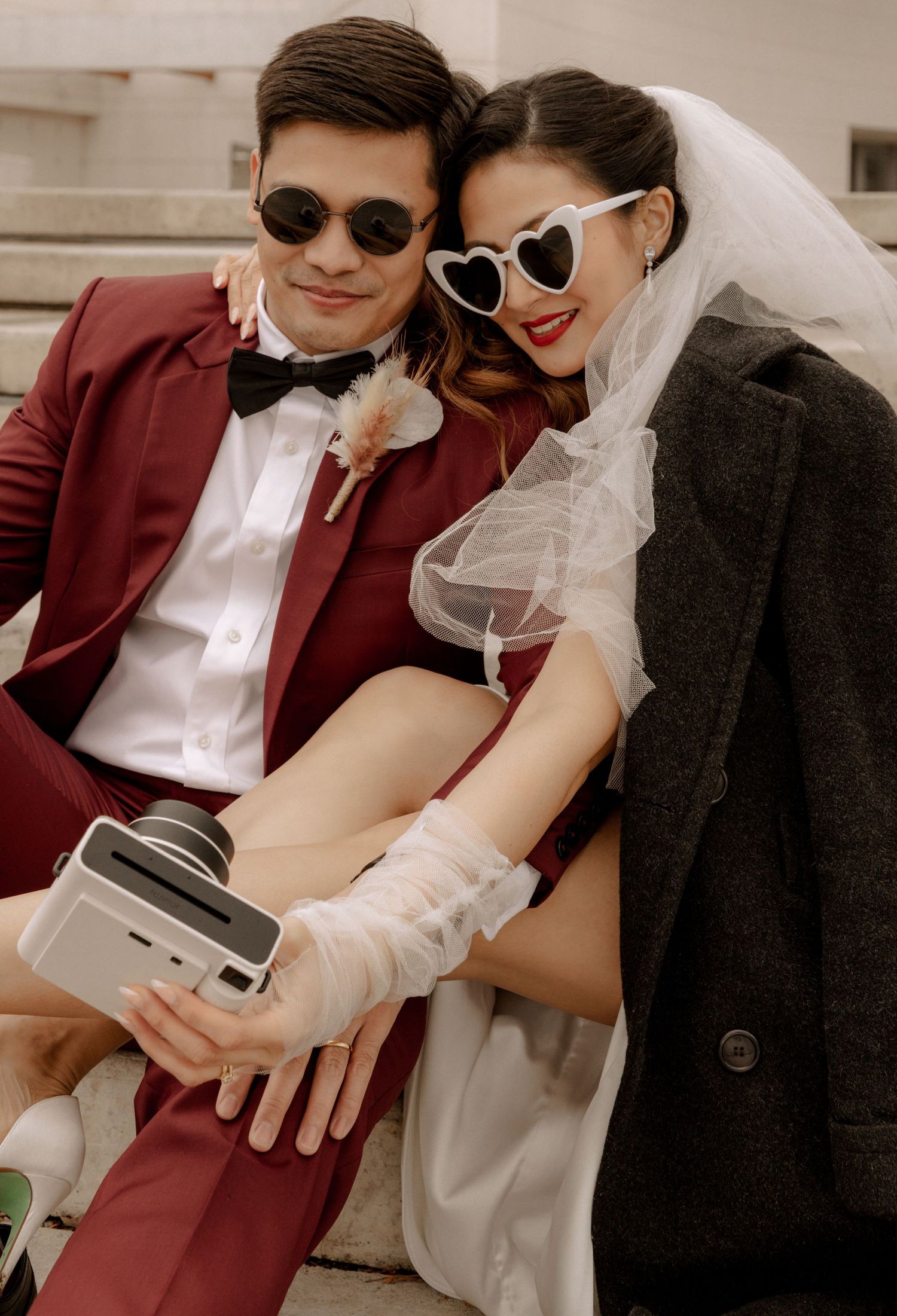 Newlyweds in retro sunglasses share playful moment with vintage camera on concrete steps.