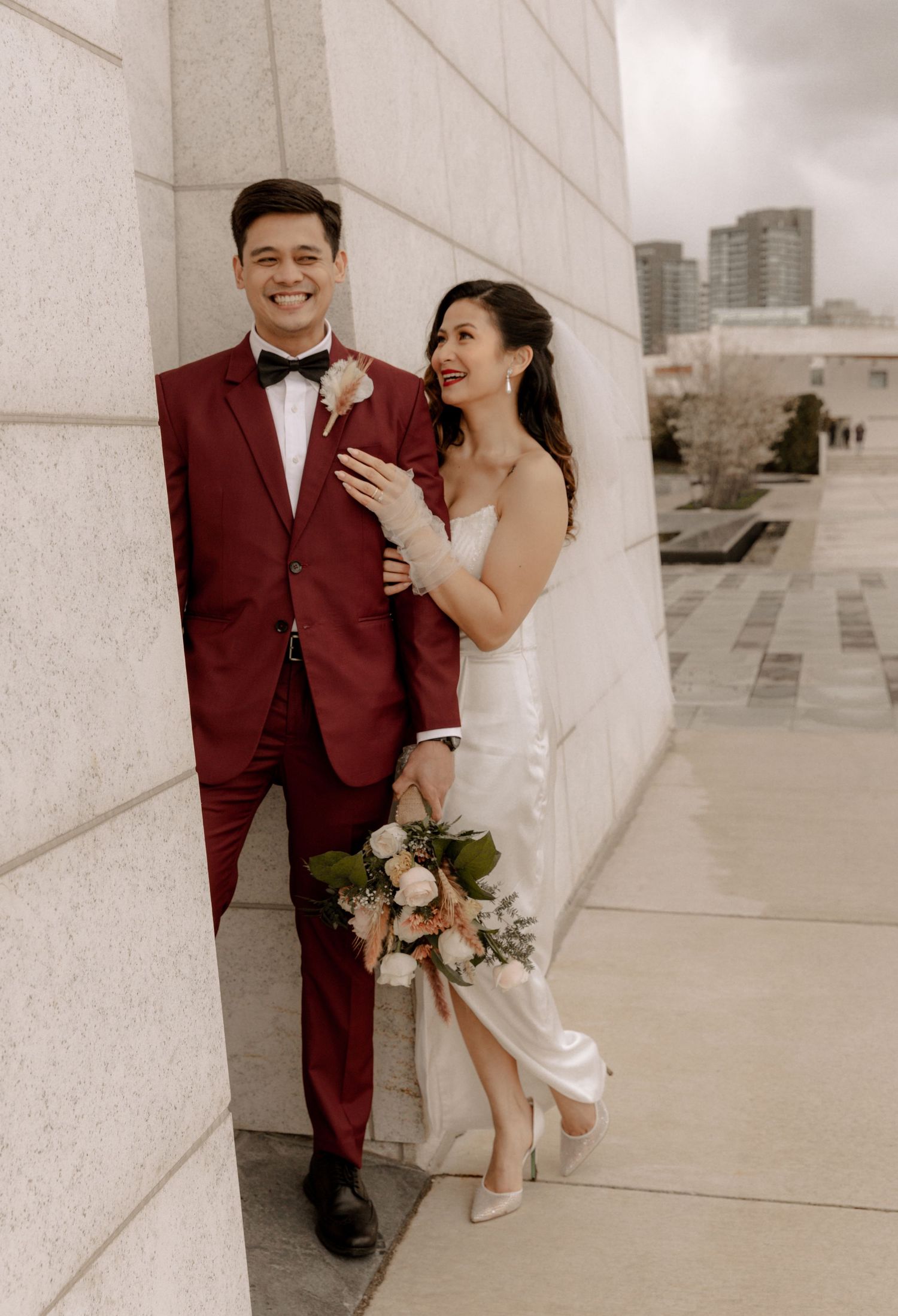 A couple poses against a white wall on their wedding day, with the bride in white and groom in burgundy suit.