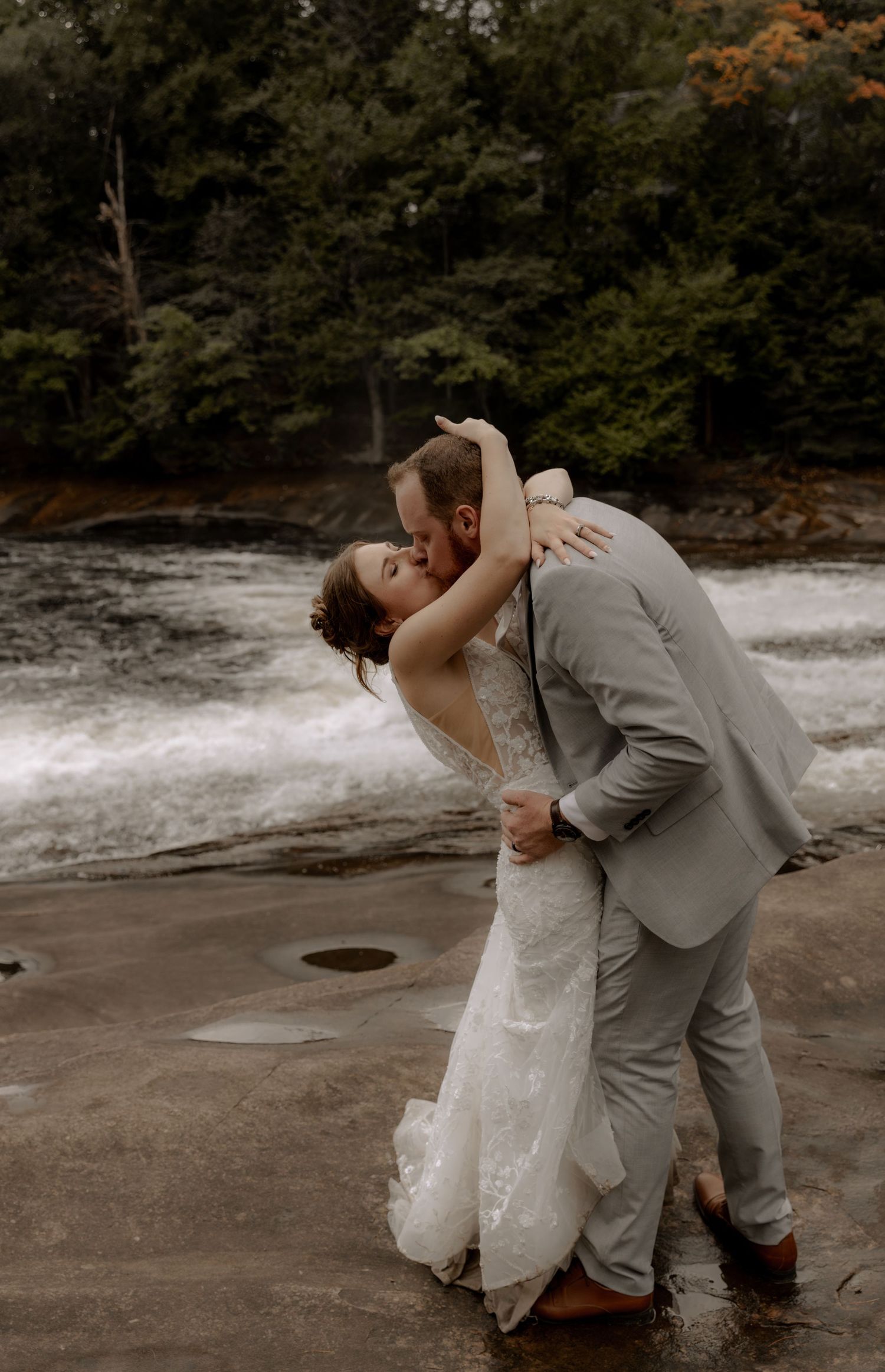 Romantic wedding dip pose on wet rocks with flowing water and forest scenery behind.