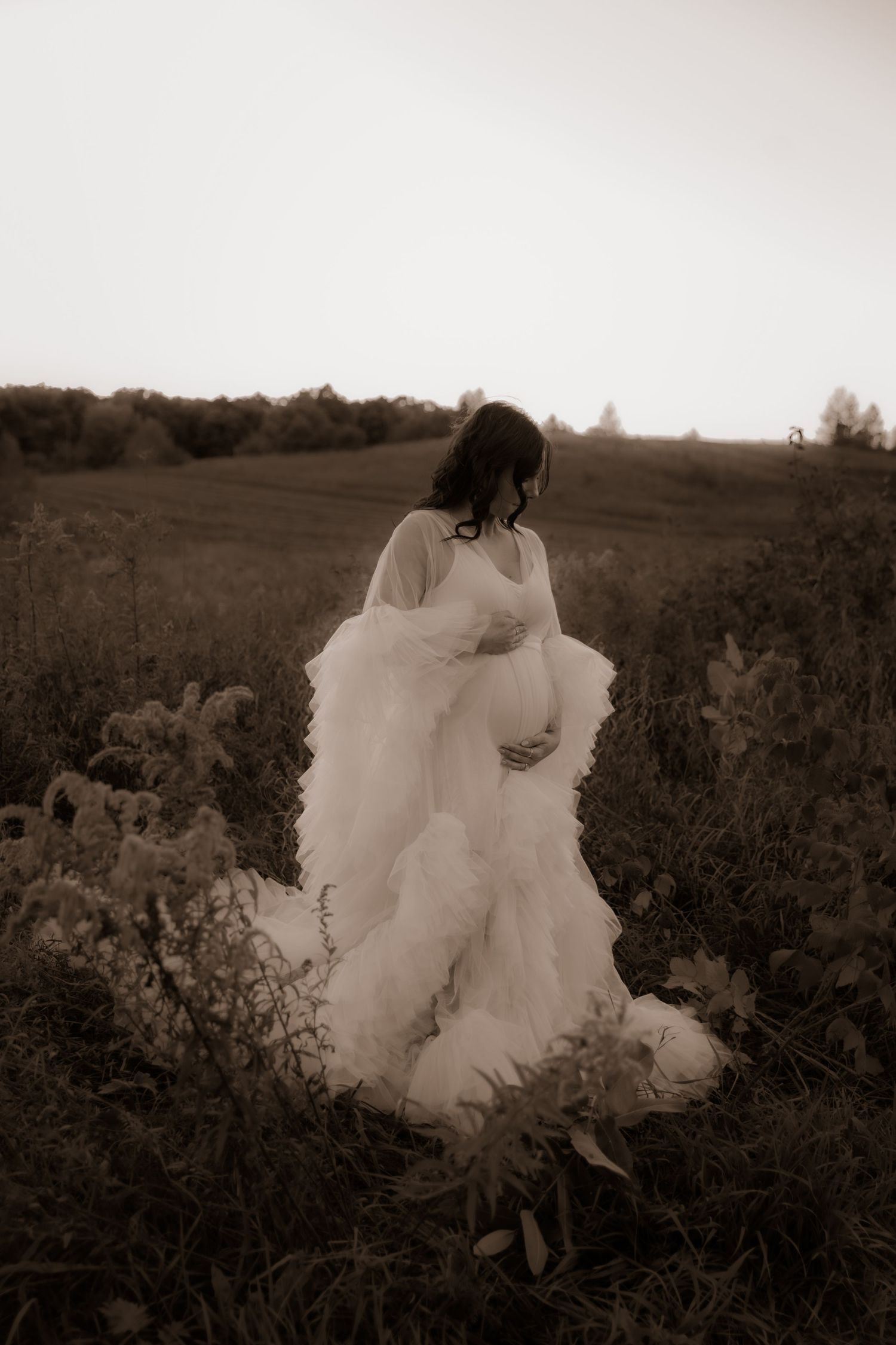A person in a flowing white wedding dress stands in a sepia-toned field of wildflowers at dusk.