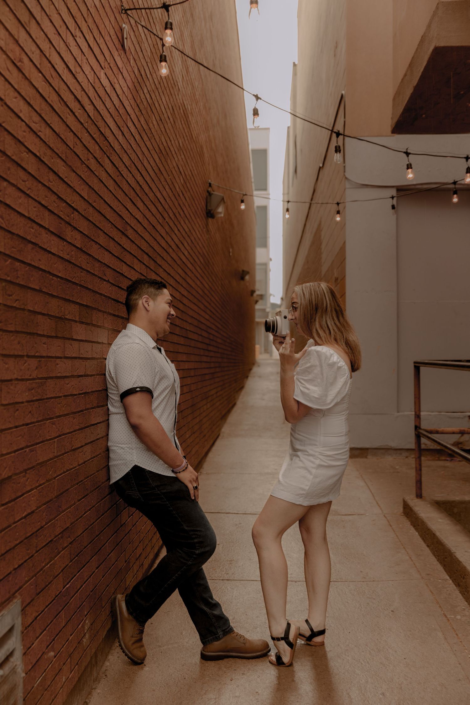 A couple standing in a narrow alley with string lights overhead and a brick wall to one side in warm moody lighting.
