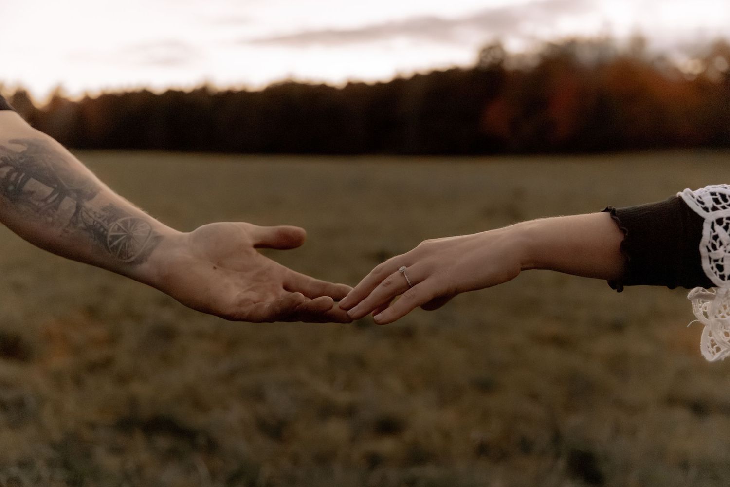 Tattooed hand reaching out to touch another hand with lace sleeve in autumn field.