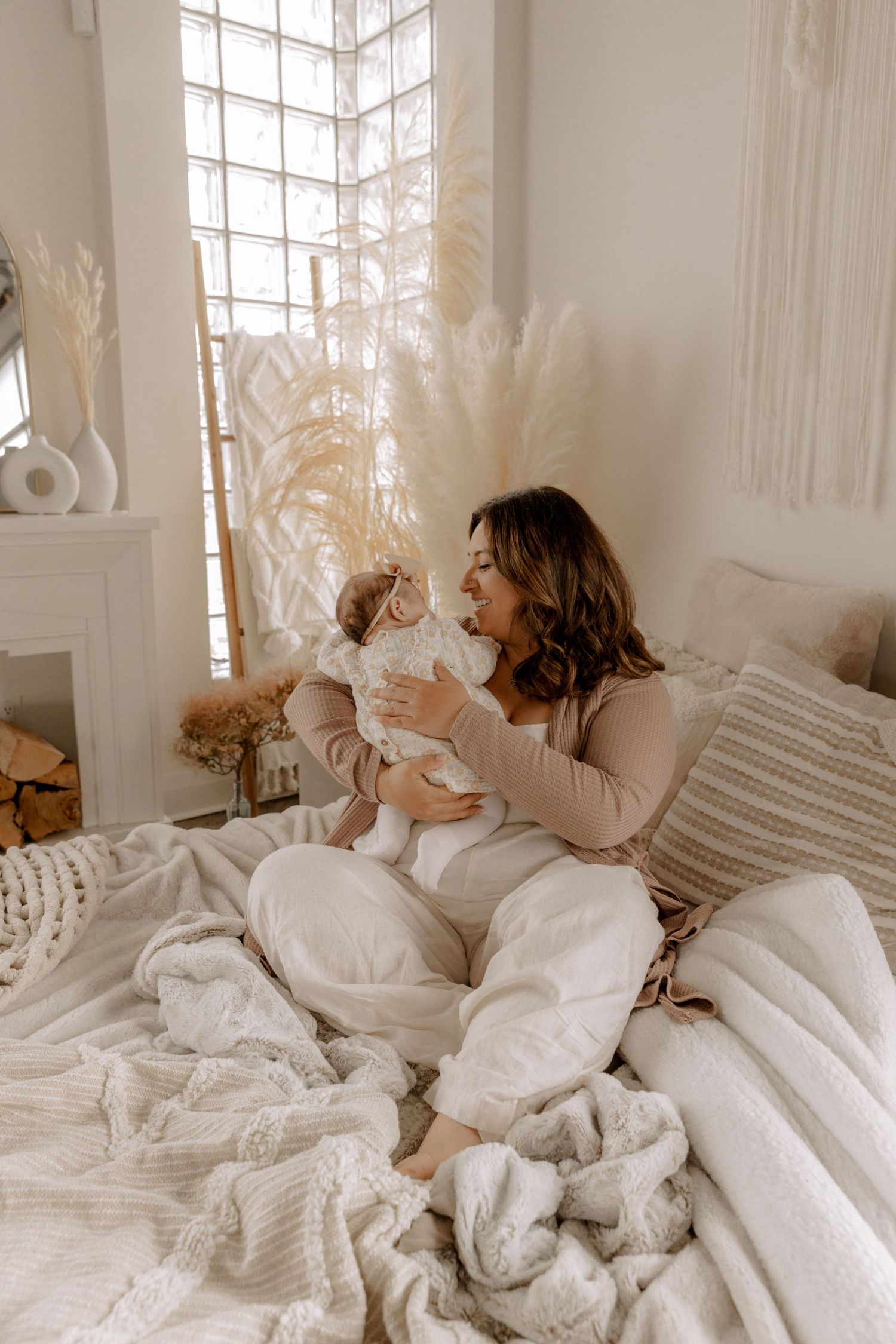 Cozy bedroom scene with white bedding and pampas grass decor as woman holds baby in soft natural window light.