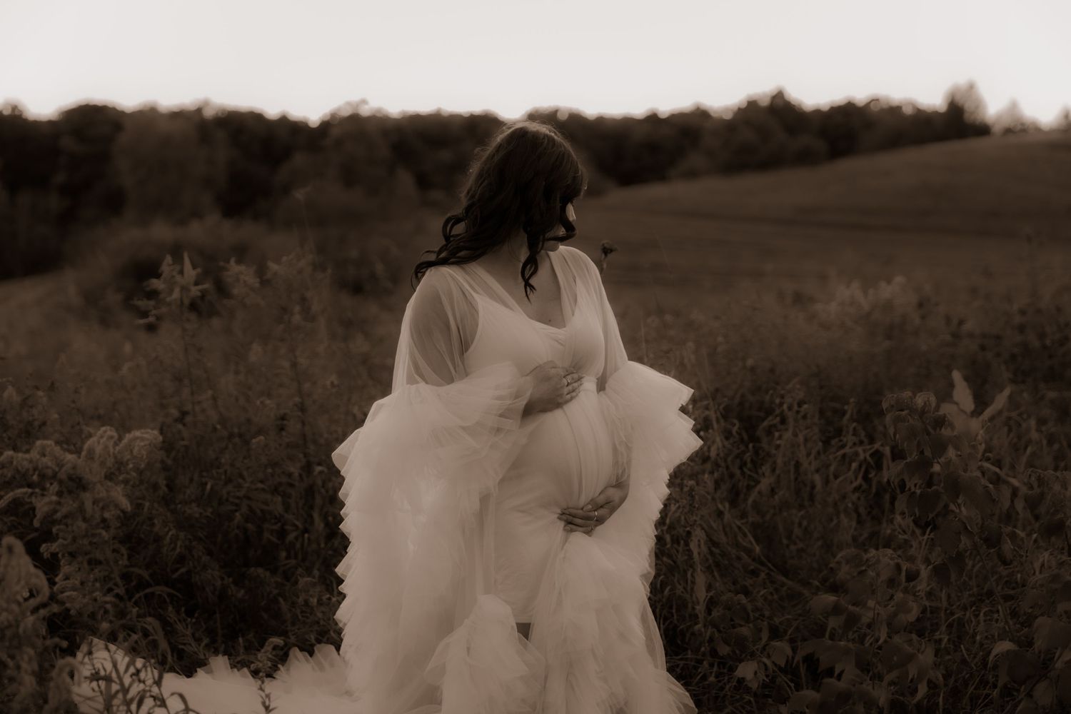 A sepia maternity photograph with a billowing white gown in a rustic field setting.