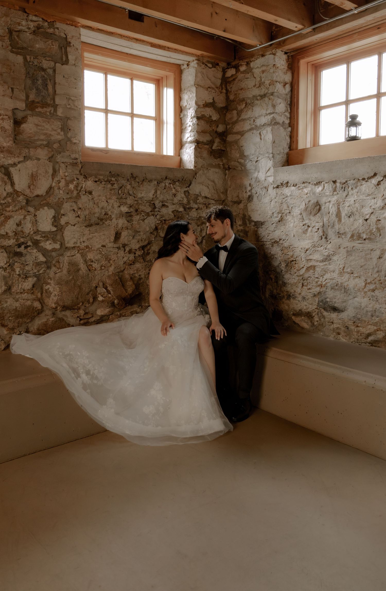 Romantic wedding photo of a couple in formal attire against stone walls with natural window light.