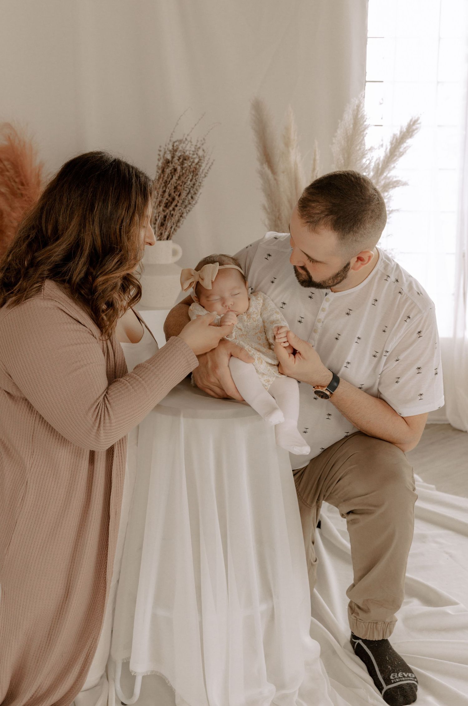 Parents in neutral tones share intimate moment with their infant surrounded by pampas grass decor.