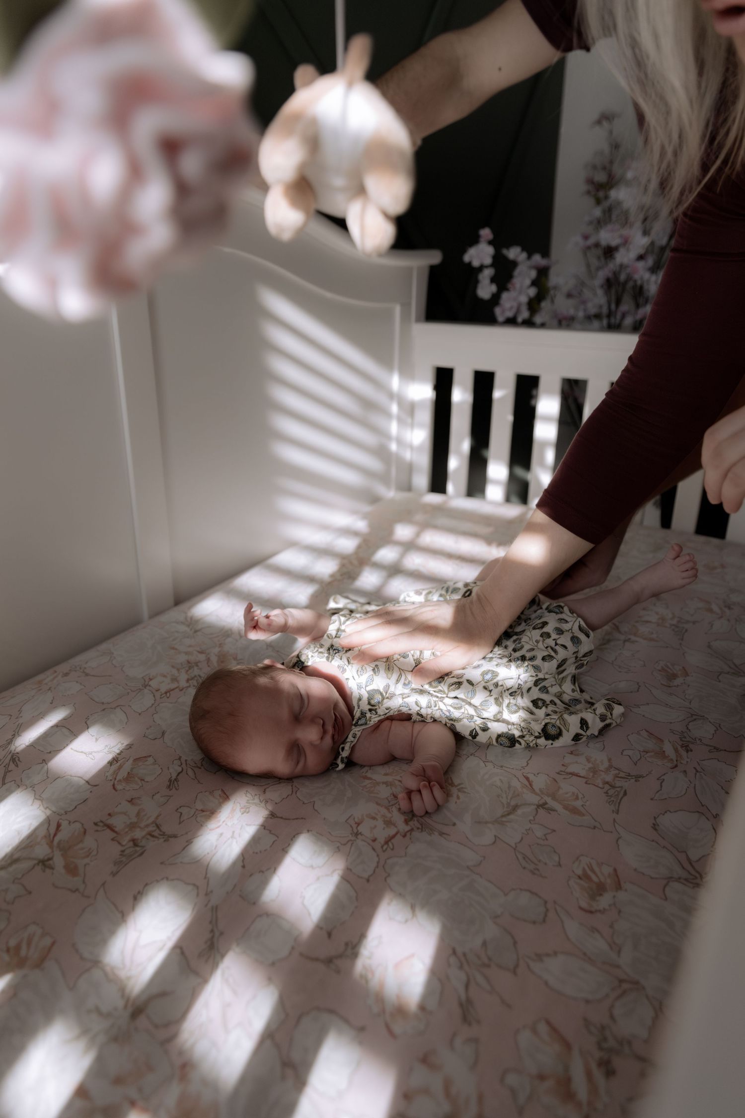 Gentle hands tend to an infant in a crib while soft light streams through window blinds.