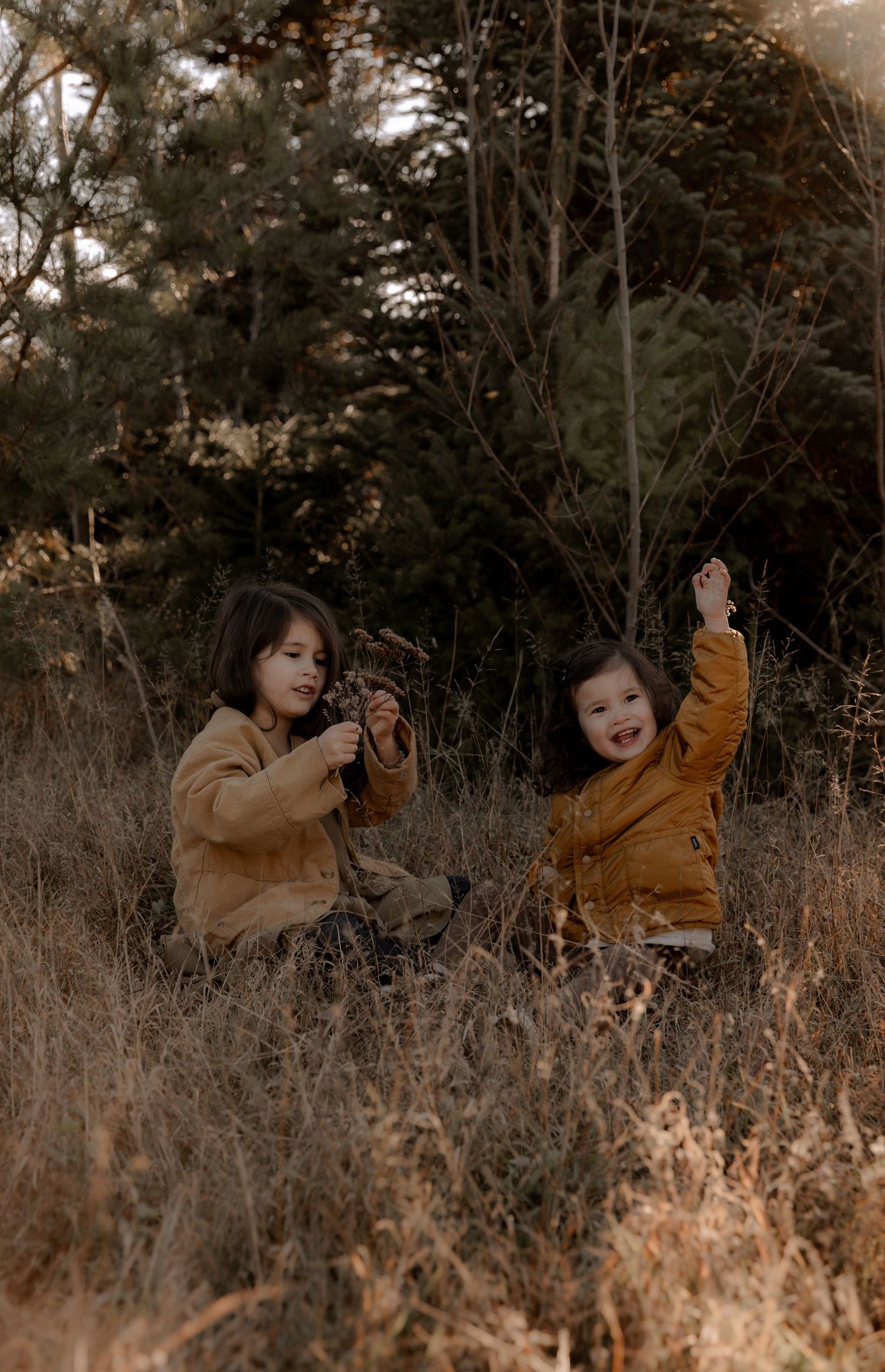 Two children in autumn outerwear playing together in a field of tall brown grass with trees behind them.