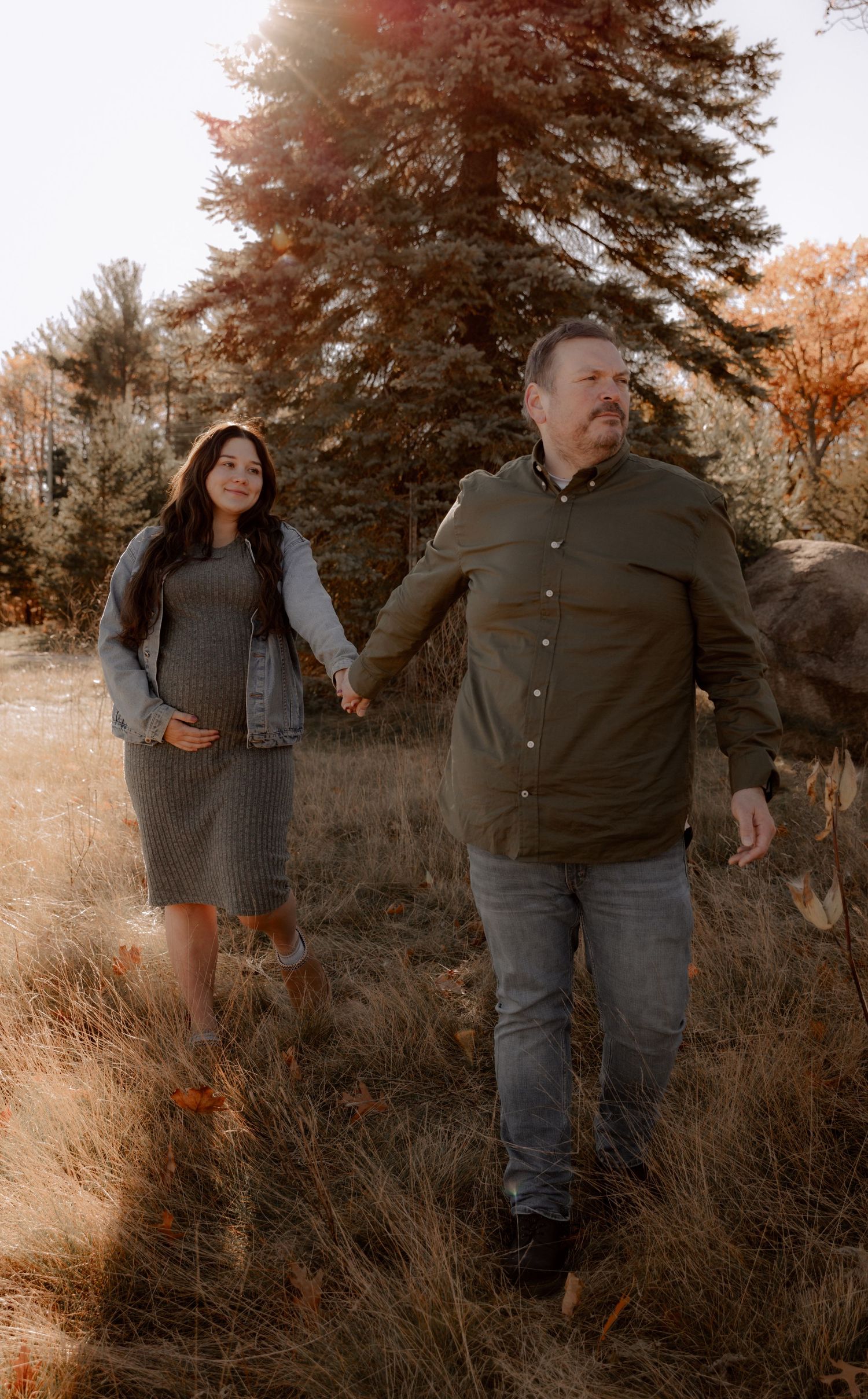 A couple holds hands while walking through tall autumn grass during golden hour at sunset in a rustic outdoor setting.
