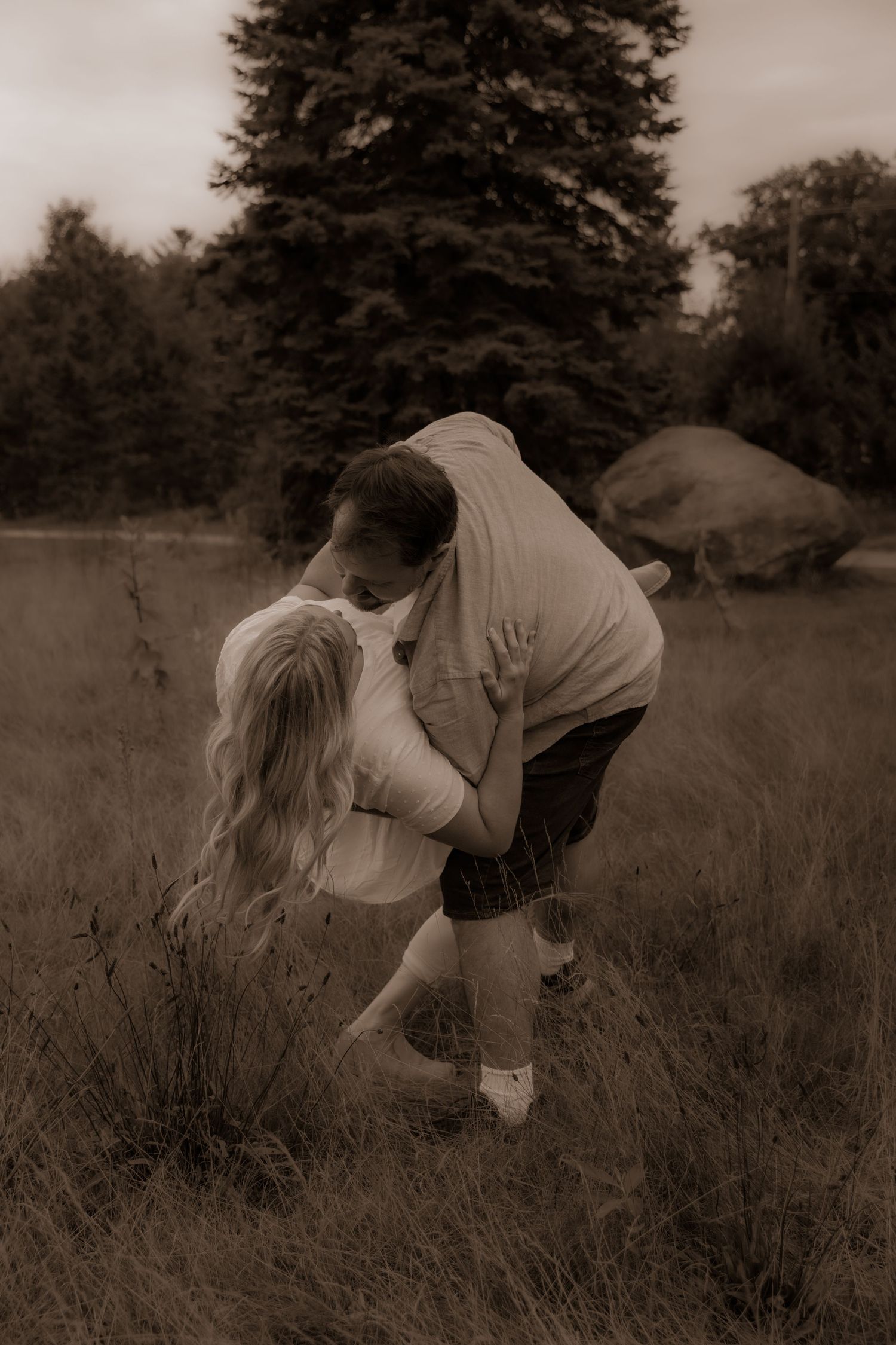 A sepia-toned romantic moment between two people in casual wear near a large evergreen tree.