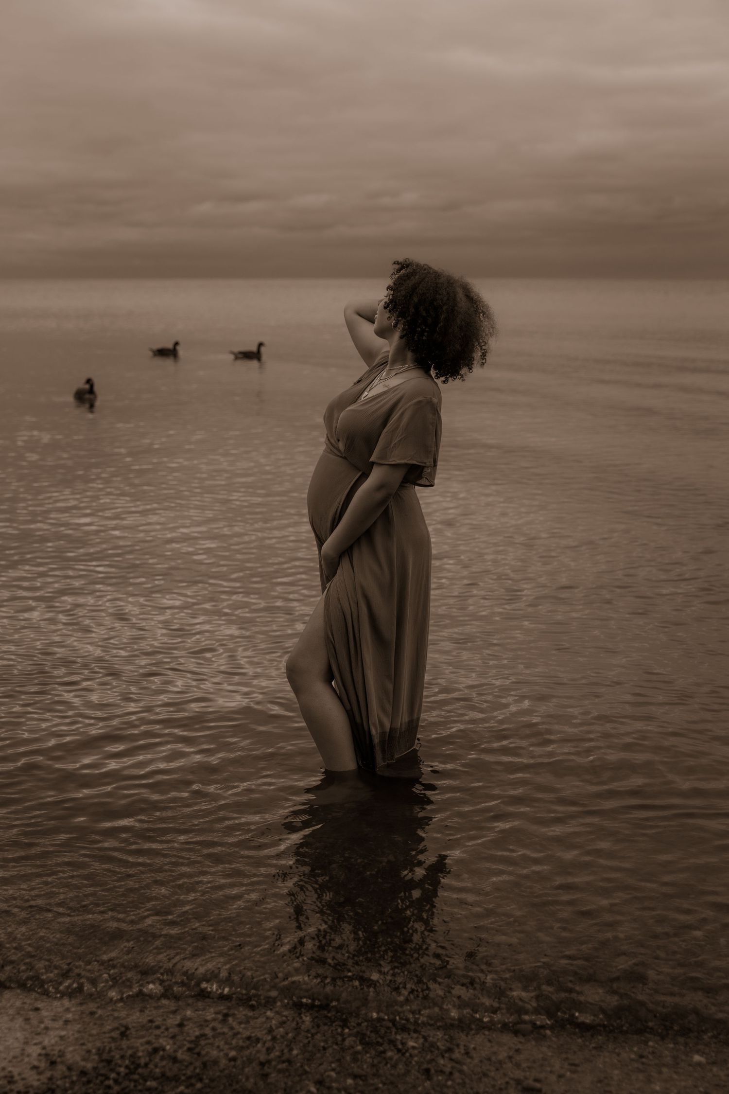 Silhouette of an expectant mother standing in shallow water at dusk with ducks swimming in the distance.