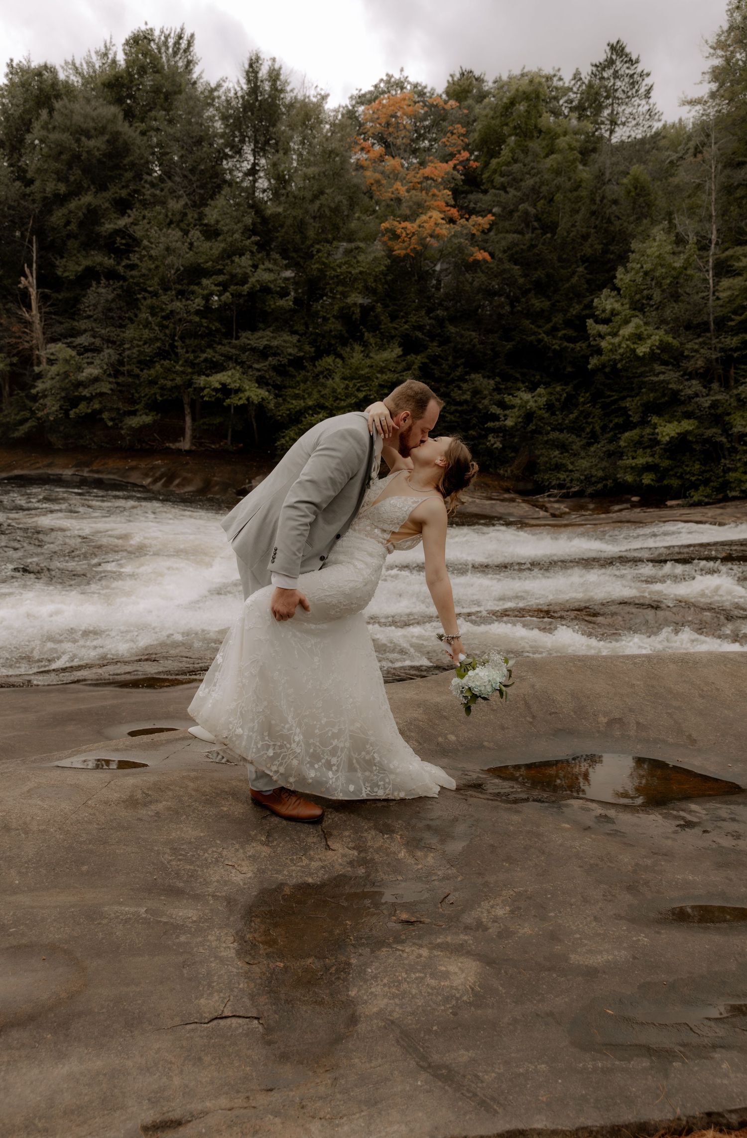 Romantic wedding dip pose on rocky waterfall shore with autumn forest backdrop.