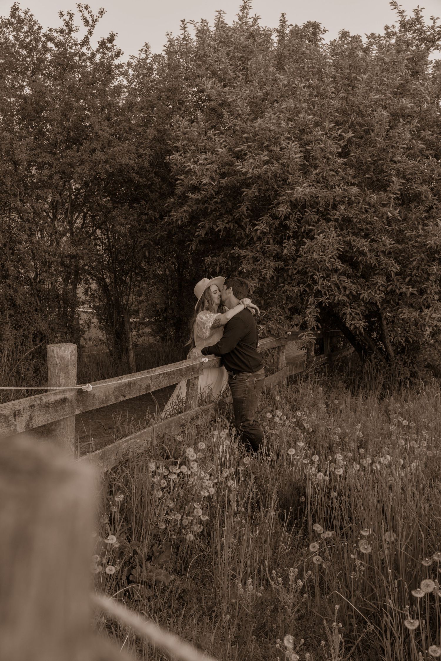 Sepia-toned photograph of a romantic moment by a wooden fence in a meadow.