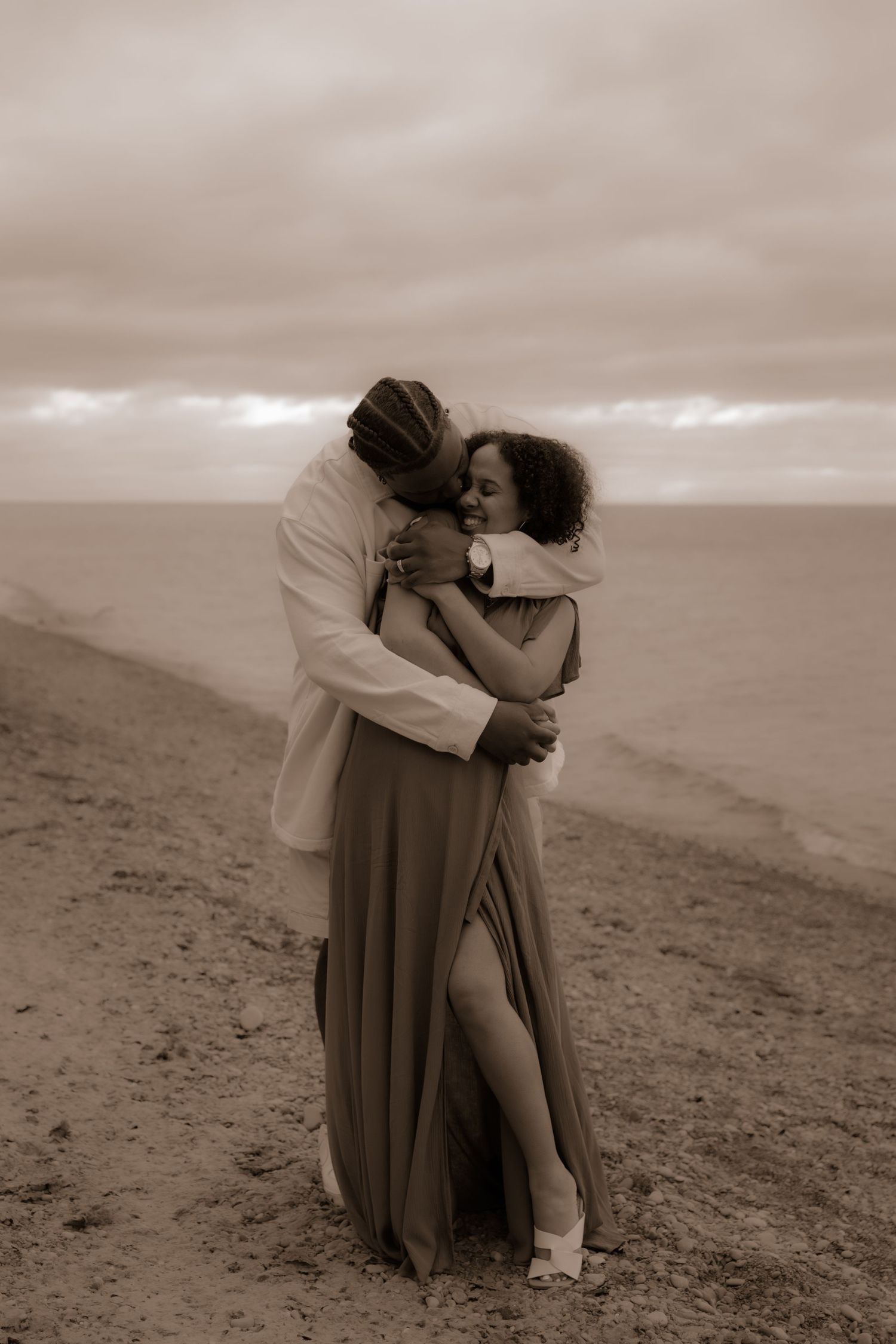 A romantic sepia-toned couple embraces on a misty beach, with flowing dress and dramatic cloudy sky backdrop.