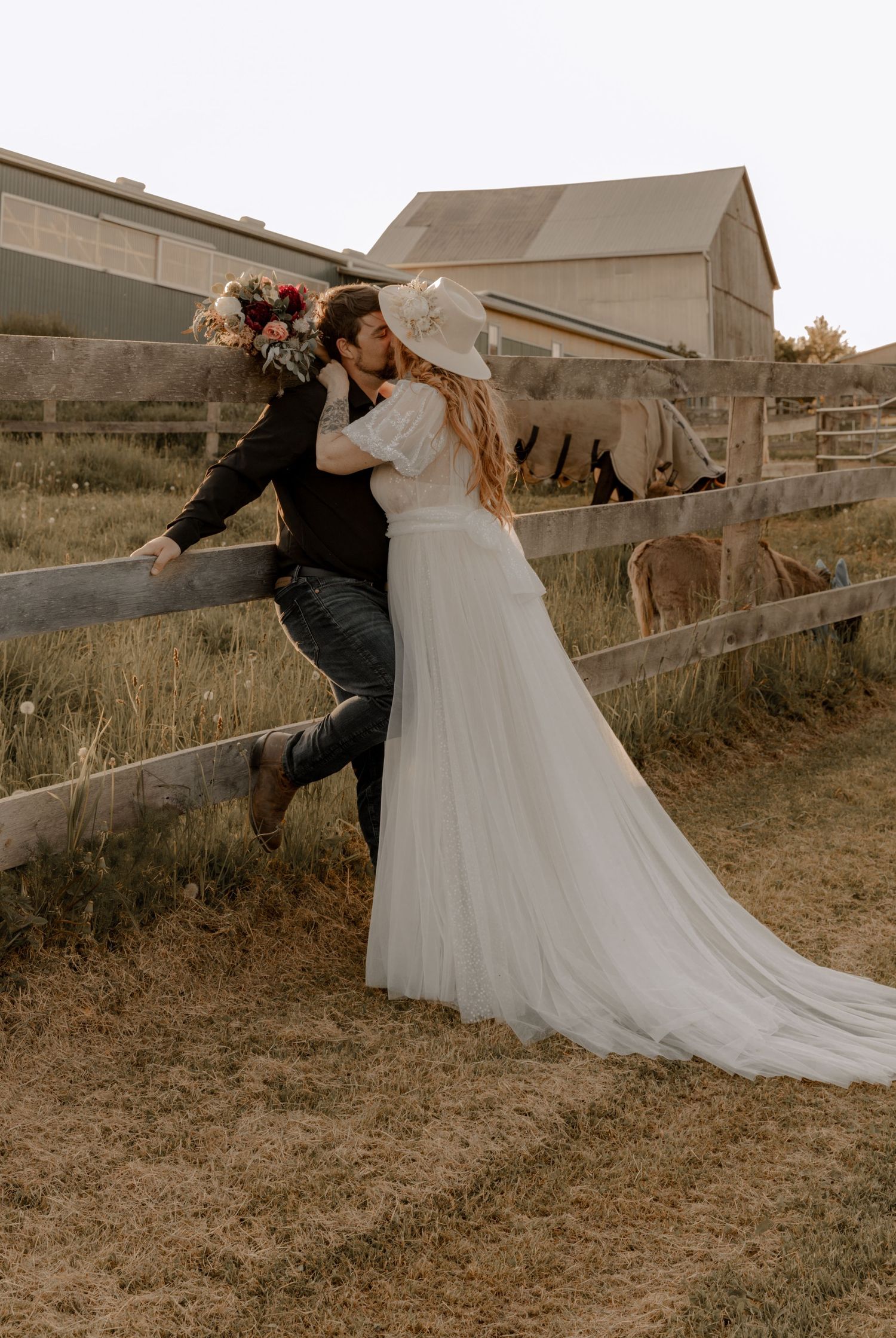 Playful wedding photo of a couple sitting on a wooden fence by a rustic barn.