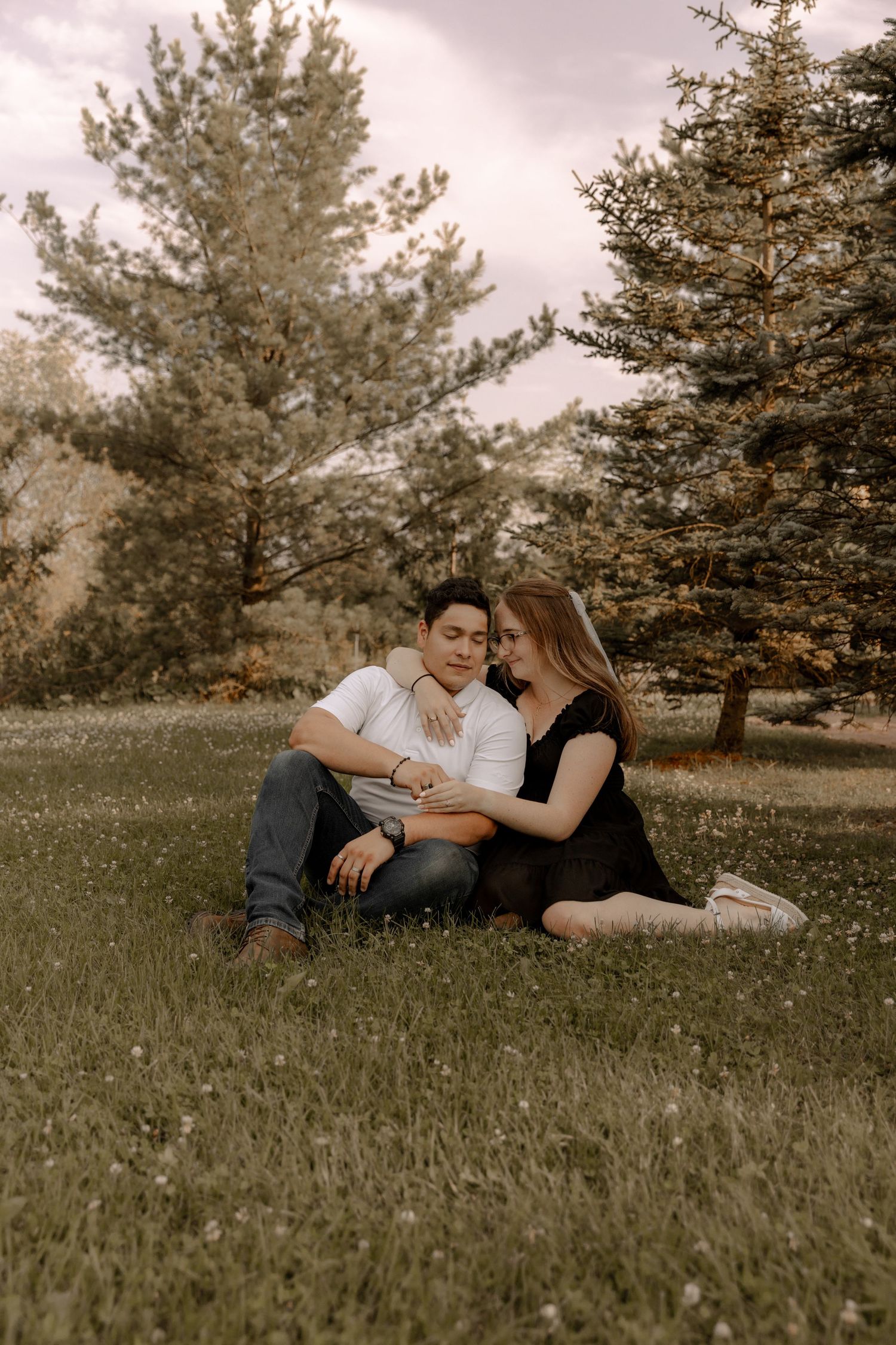 A couple sits intimately on grass surrounded by evergreen trees at sunset.