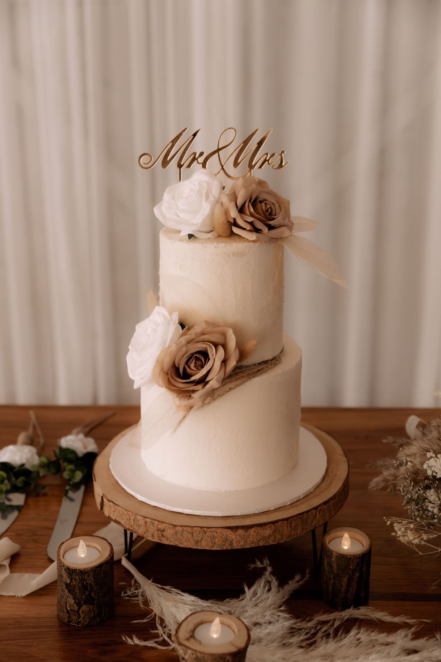 Elegant two-tier white wedding cake with brown roses and 'Mr & Mrs' topper surrounded by candles.