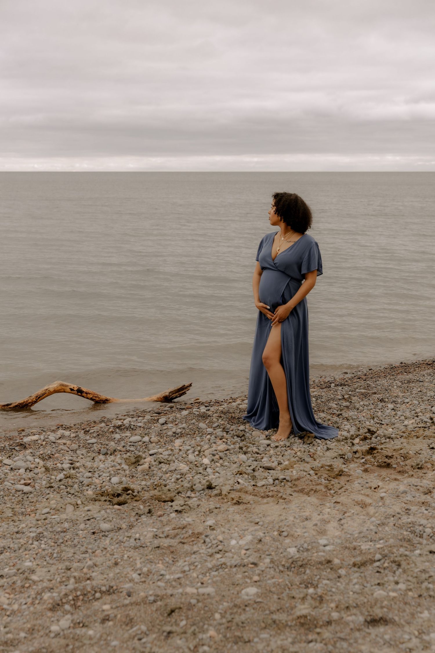 A pregnant person in a flowing blue dress stands on a rocky beach shore against a moody grey sky.