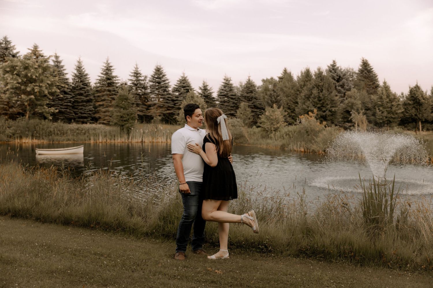 A couple embraces on a grassy path beside a tranquil pond with evergreen trees in the background.