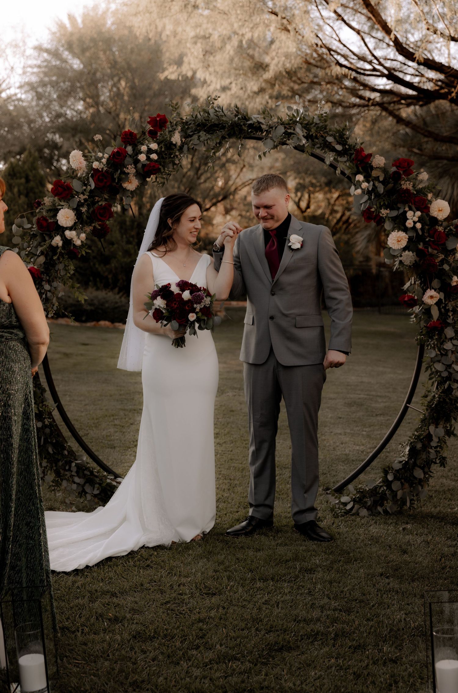 A wedding couple stands under a floral arch with burgundy and blush blooms in a garden setting.